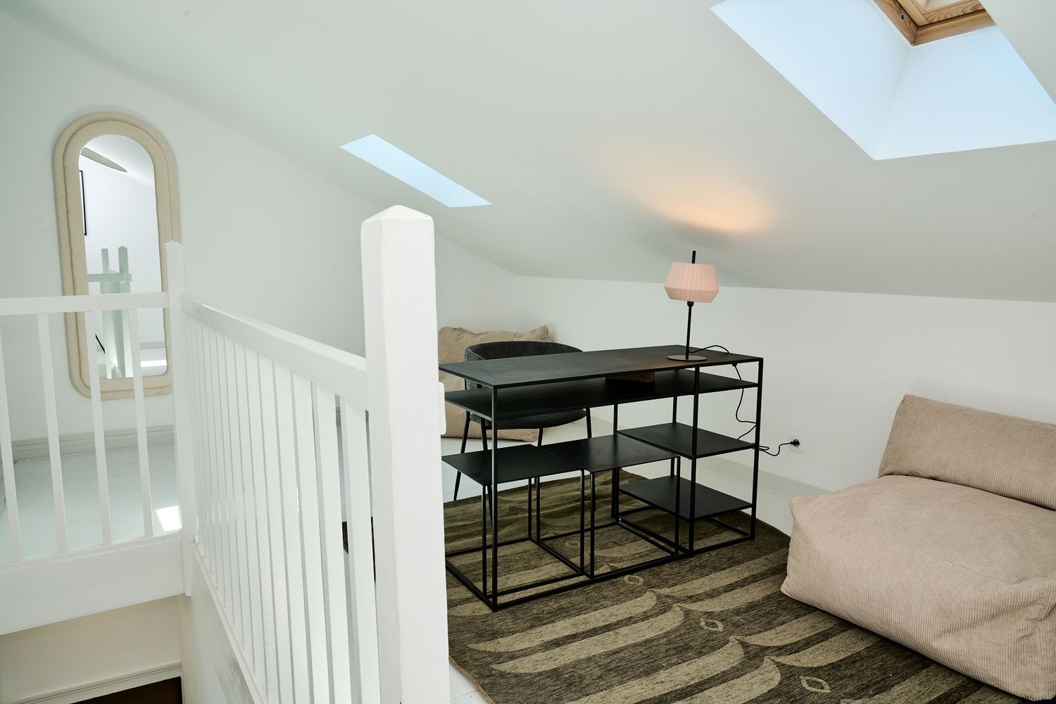 Attic loft space with skylight, black metal shelving unit, sofa, and arched mirror at top of white staircase.