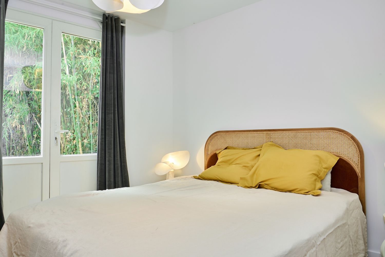 Bright minimalist bedroom with rattan headboard, yellow pillows, white bedding, and a large window with green garden views.