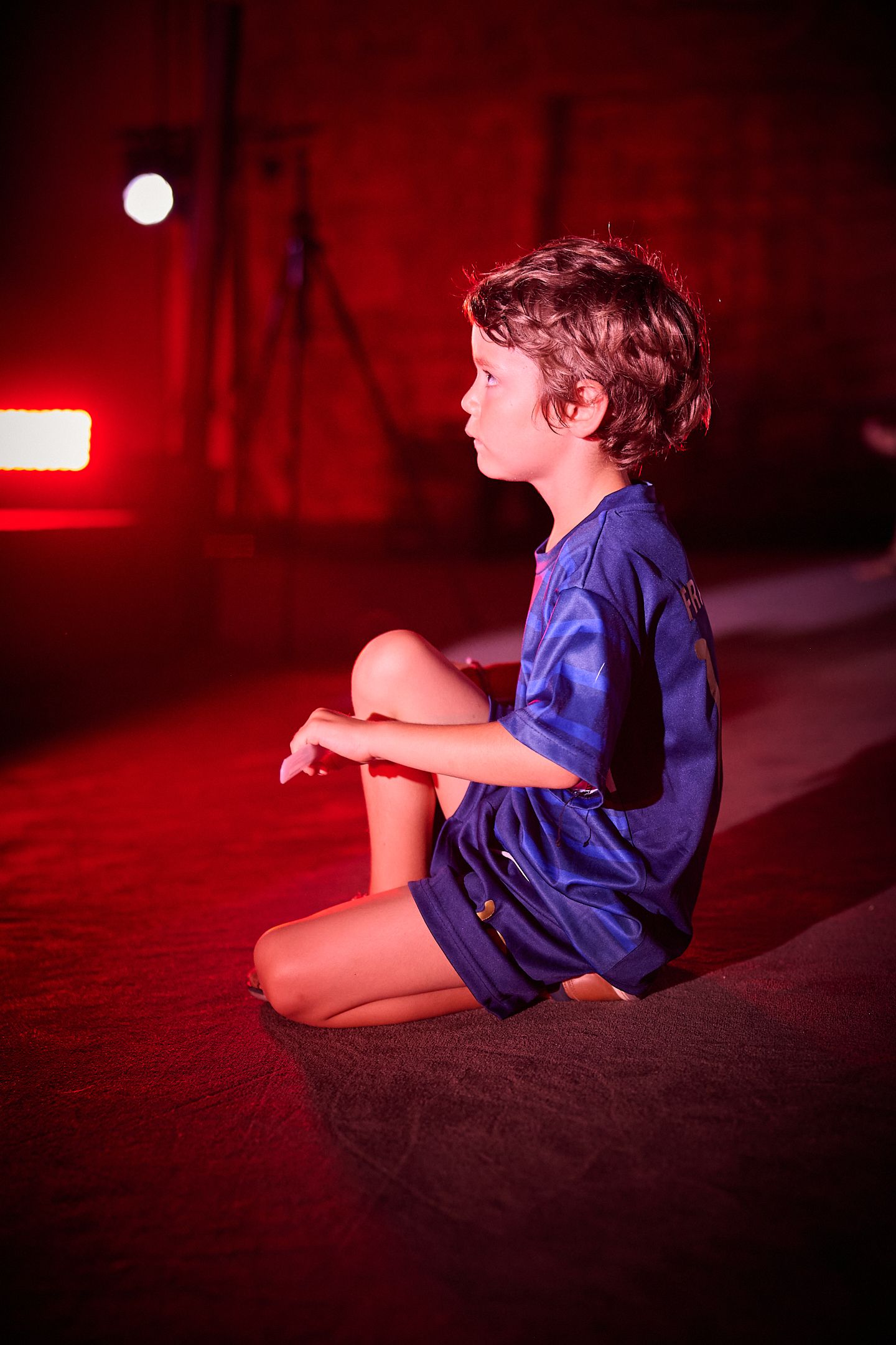 A young boy in blue shorts sits cross-legged on a stage floor, illuminated by red and white dramatic lighting.
