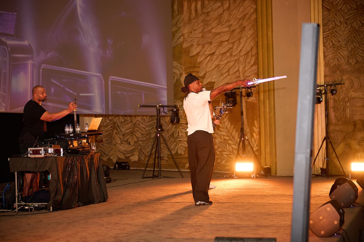 Performer wielding glowing light sabers on stage while a musician plays keyboard in a dramatic theater setting.