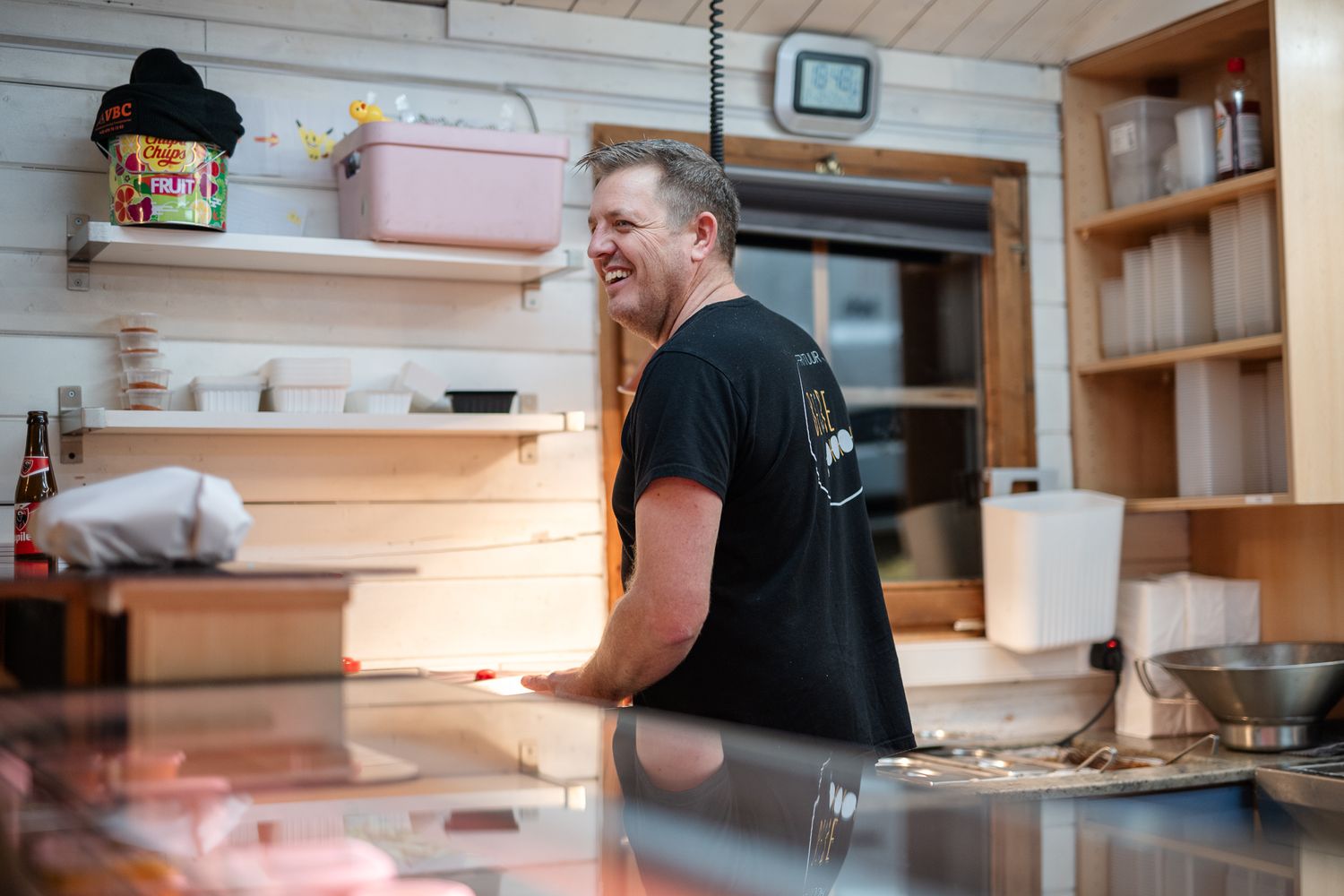 Smiling man in black t-shirt standing behind counter in cozy wooden restaurant kitchen.