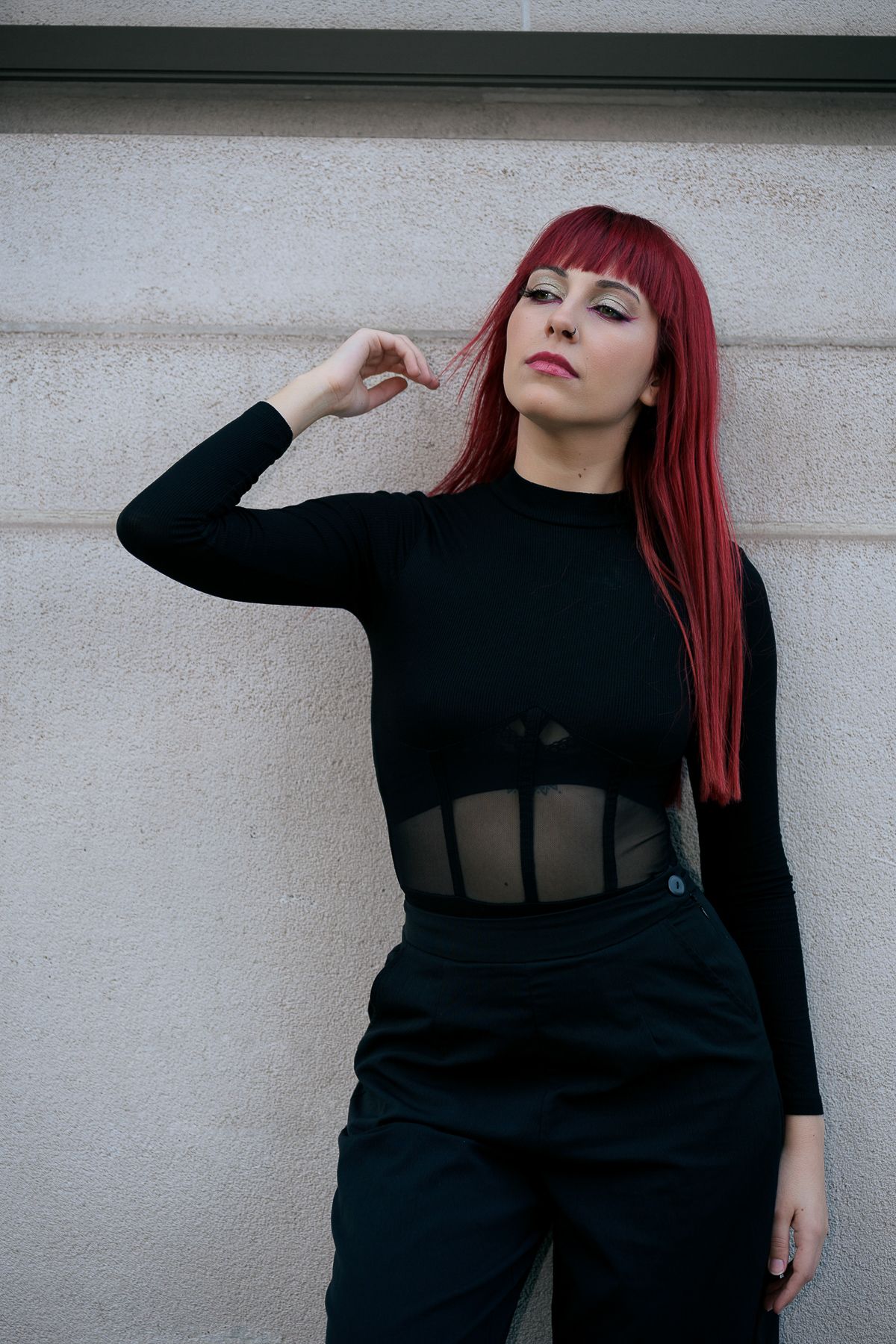 Red-haired woman in black mesh bodysuit and pants posing against a gray wall.