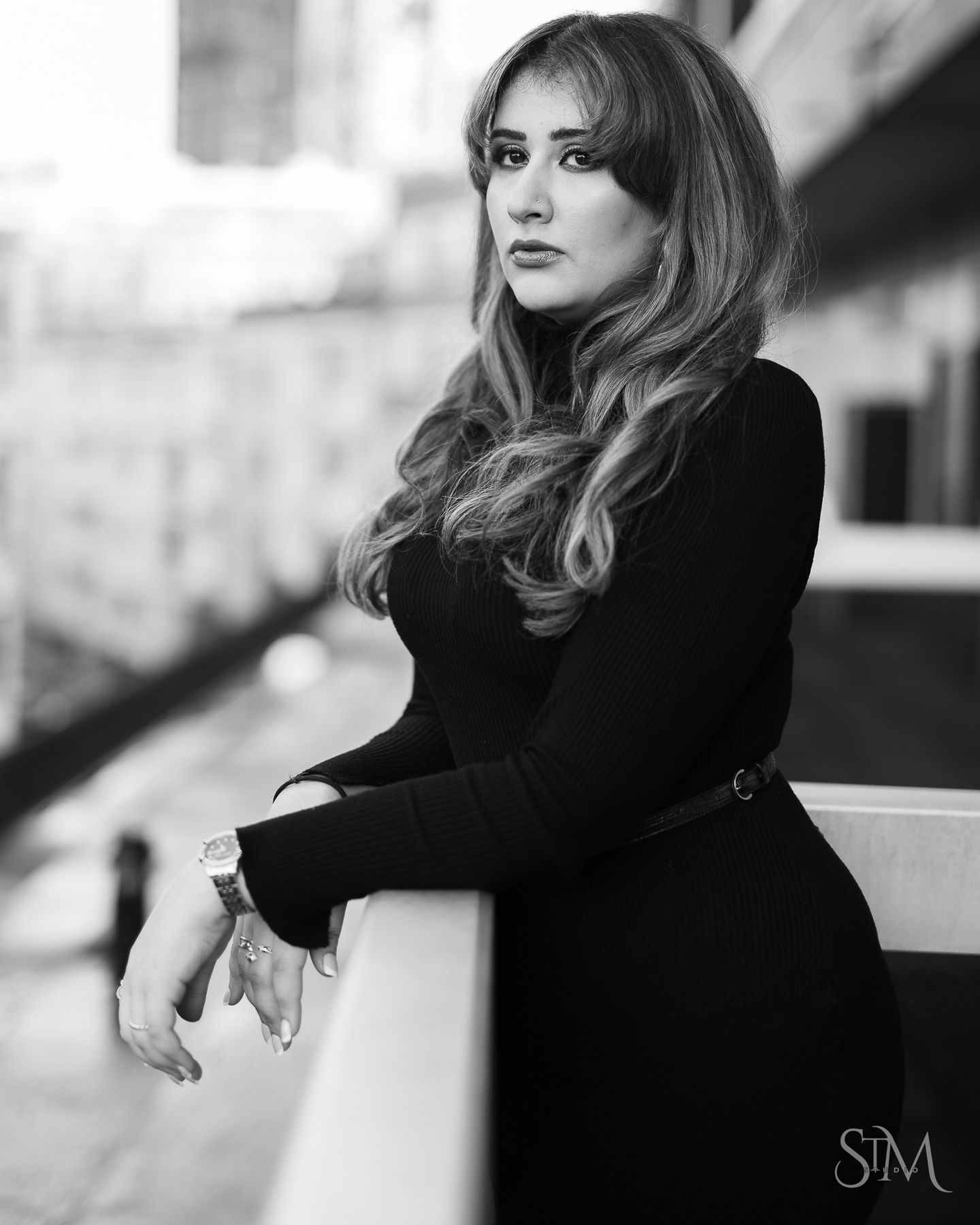 Black and white portrait of a woman in a black dress leaning on a railing with city buildings in the background.