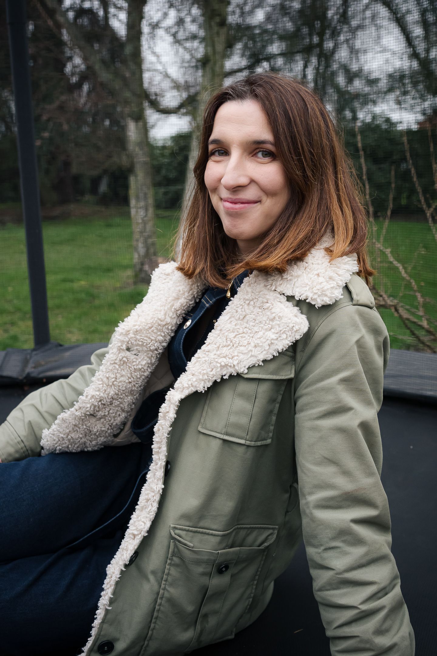 Smiling woman in olive shearling jacket sitting on a trampoline outdoors.