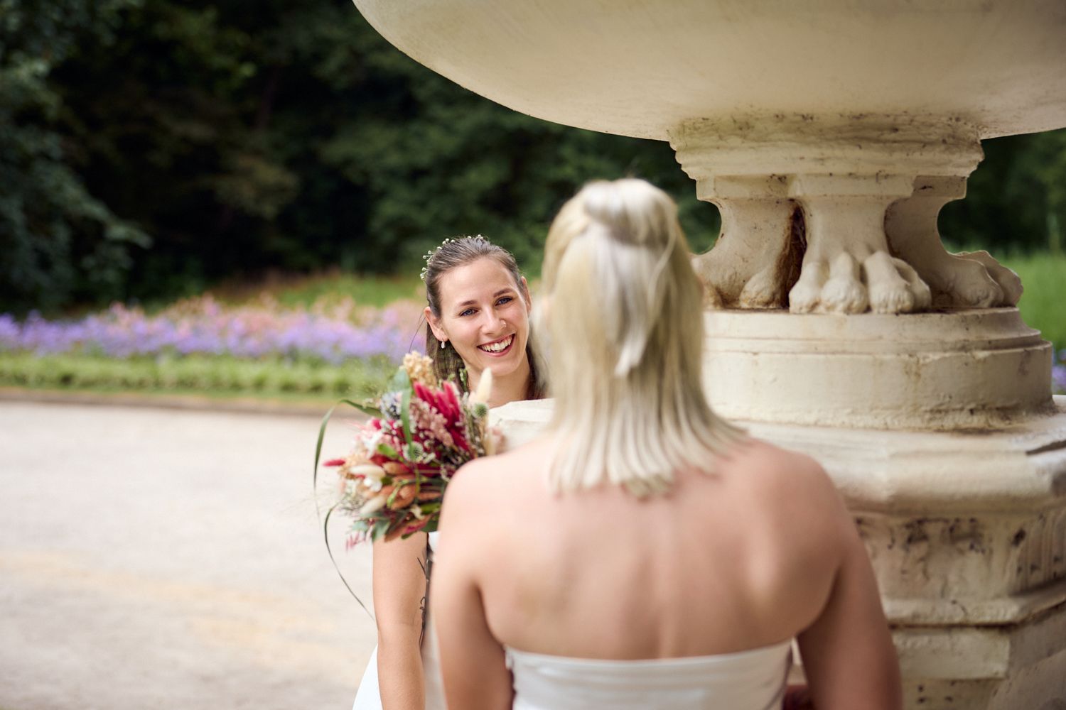 Two brides in white dresses share a joyful moment near a stone fountain, one holding a colorful bridal bouquet.