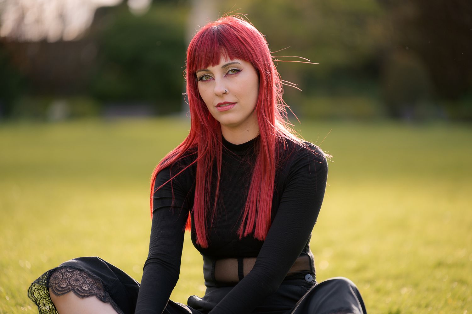 Young woman with vibrant red hair in black outfit sits on green grass in a sunny park, gazing at camera.