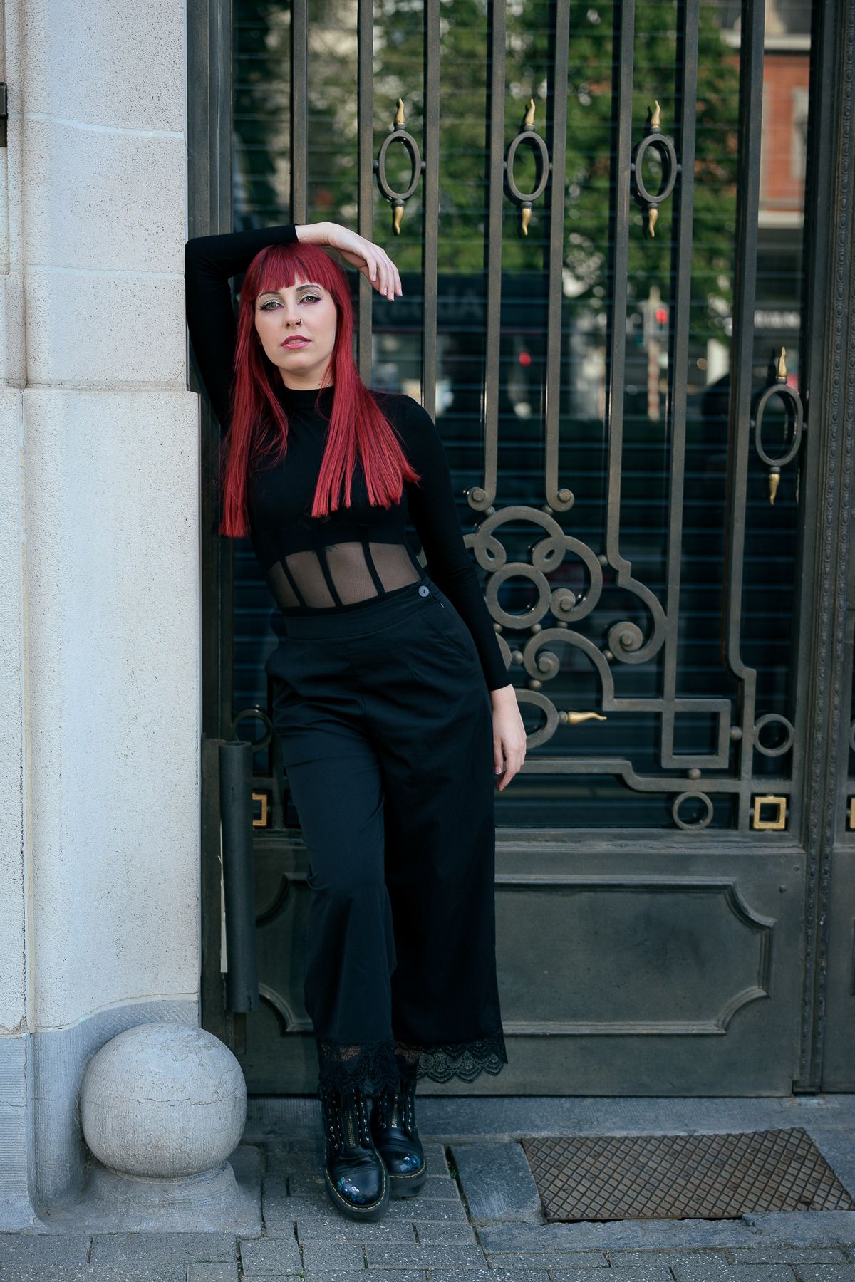 Woman with red hair in all-black outfit leaning against ornate iron gate outdoors.