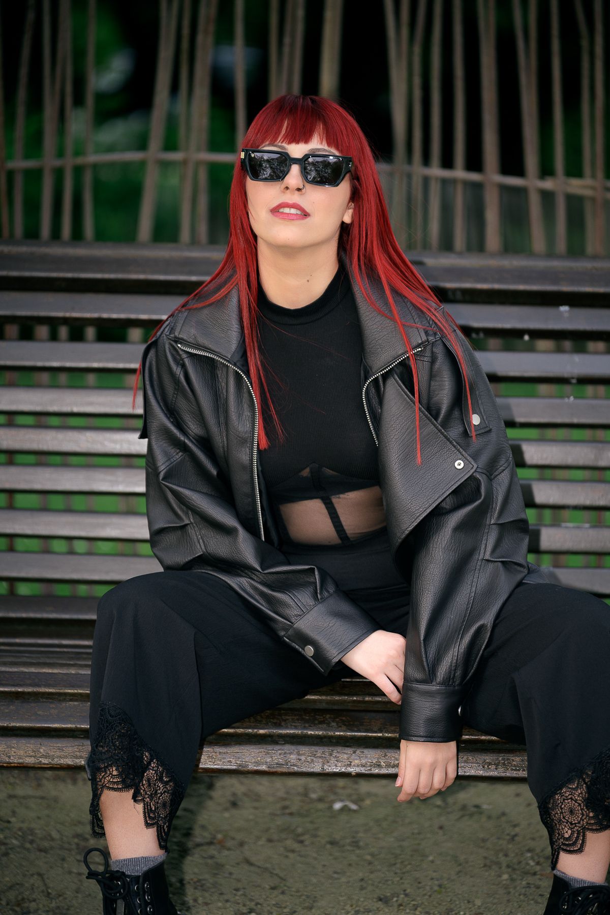Red-haired woman in black leather jacket and sunglasses sits on a bench outdoors.