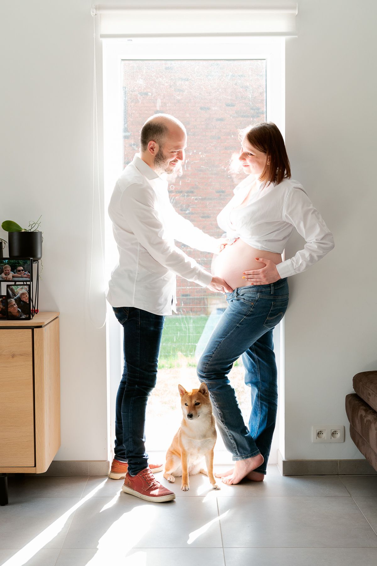 Pregnant couple in white shirts and jeans standing by window with Shiba Inu dog in bright living room.