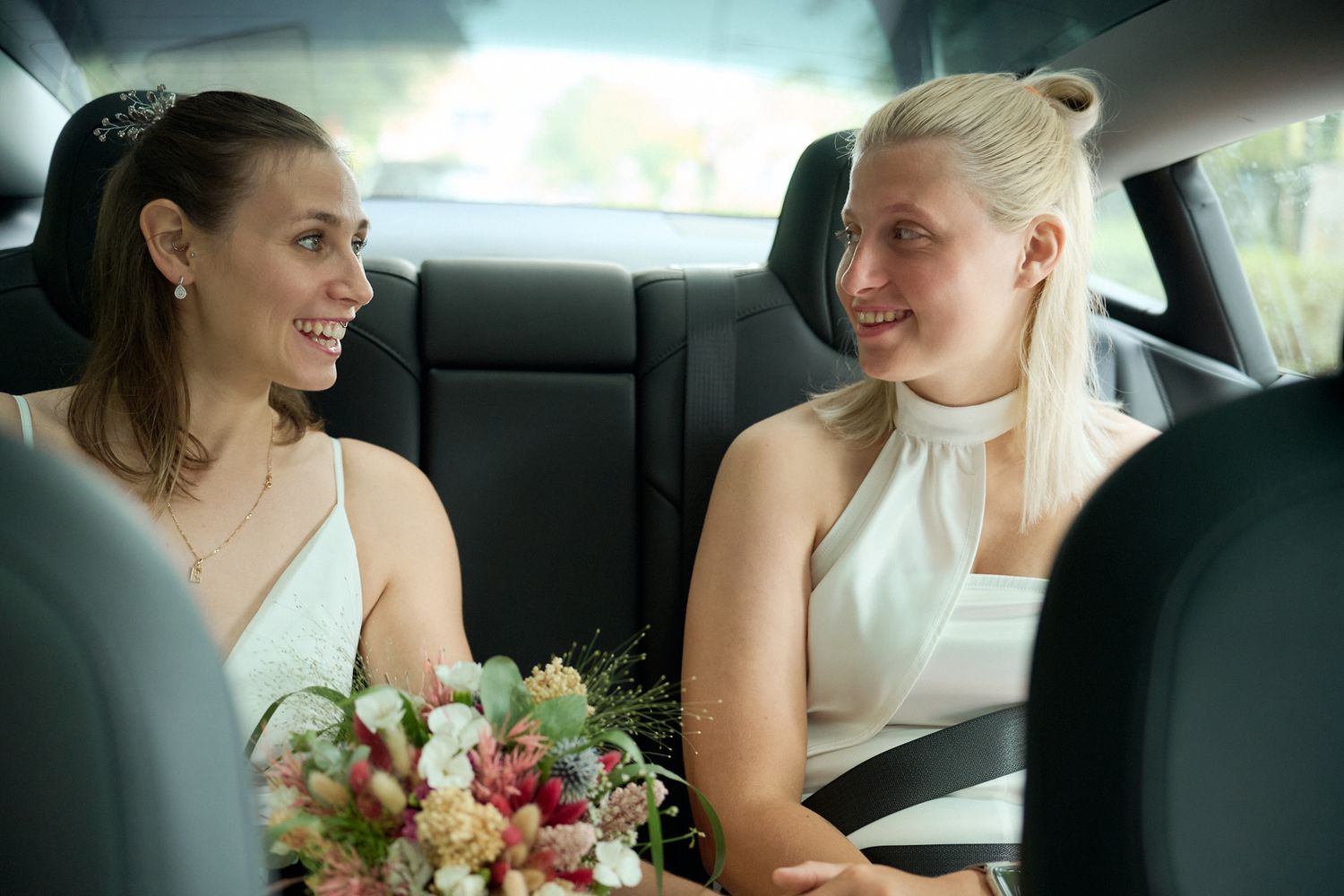 Two brides in white dresses smile at each other in a car, one holding a bouquet of flowers.