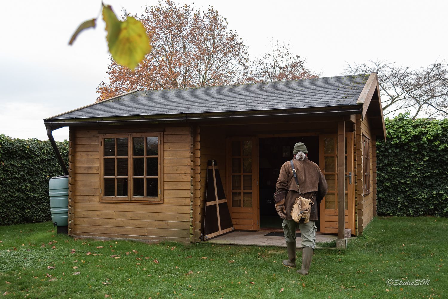Man standing outside a wooden garden cabin with large windows, surrounded by green hedges and autumn trees.