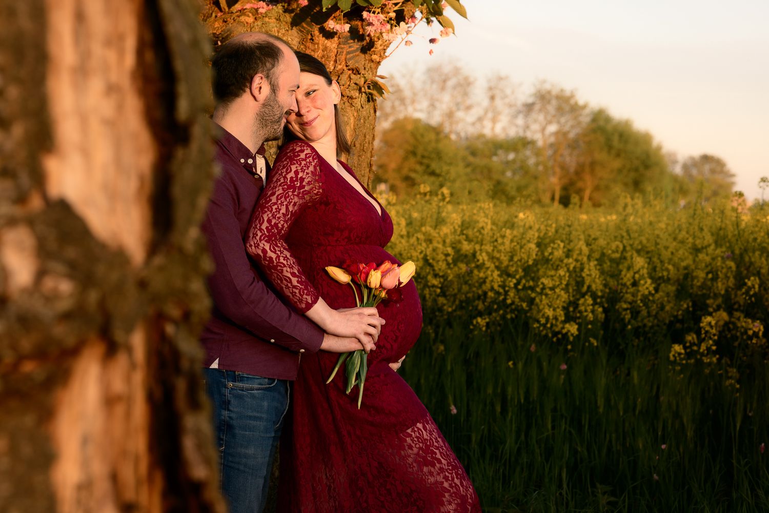 Couple sharing a tender moment outdoors, woman in red lace dress holding tulips, golden hour light, yellow flower field backdrop.