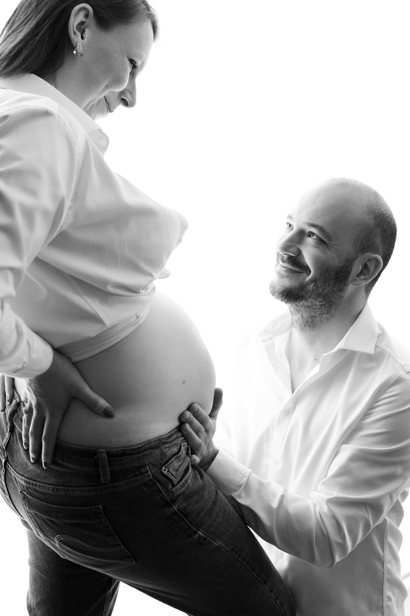 Black and white photo of a smiling pregnant couple, man kneeling beside woman's baby bump in white shirts.