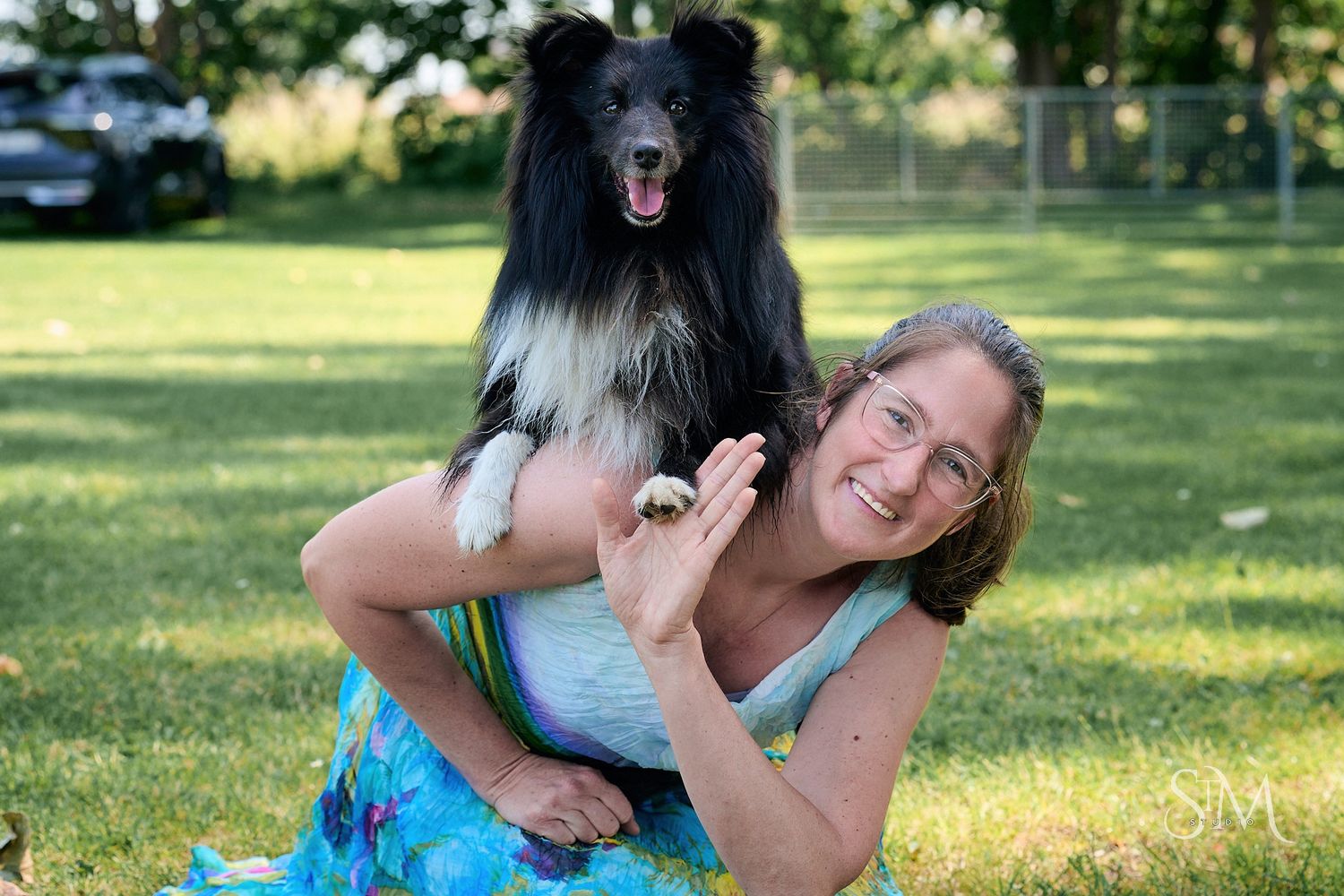 Woman smiling with a black and white Shetland Sheepdog on her back in a green park setting.