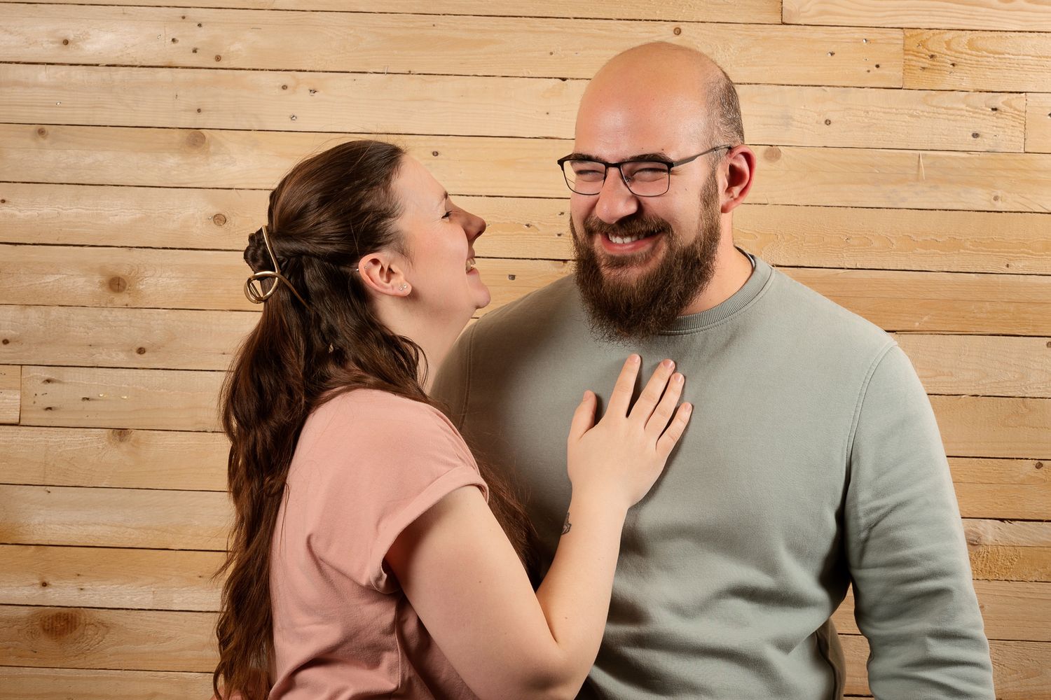 A smiling couple shares a joyful moment, woman in pink top touching bearded man's chest against wood wall.