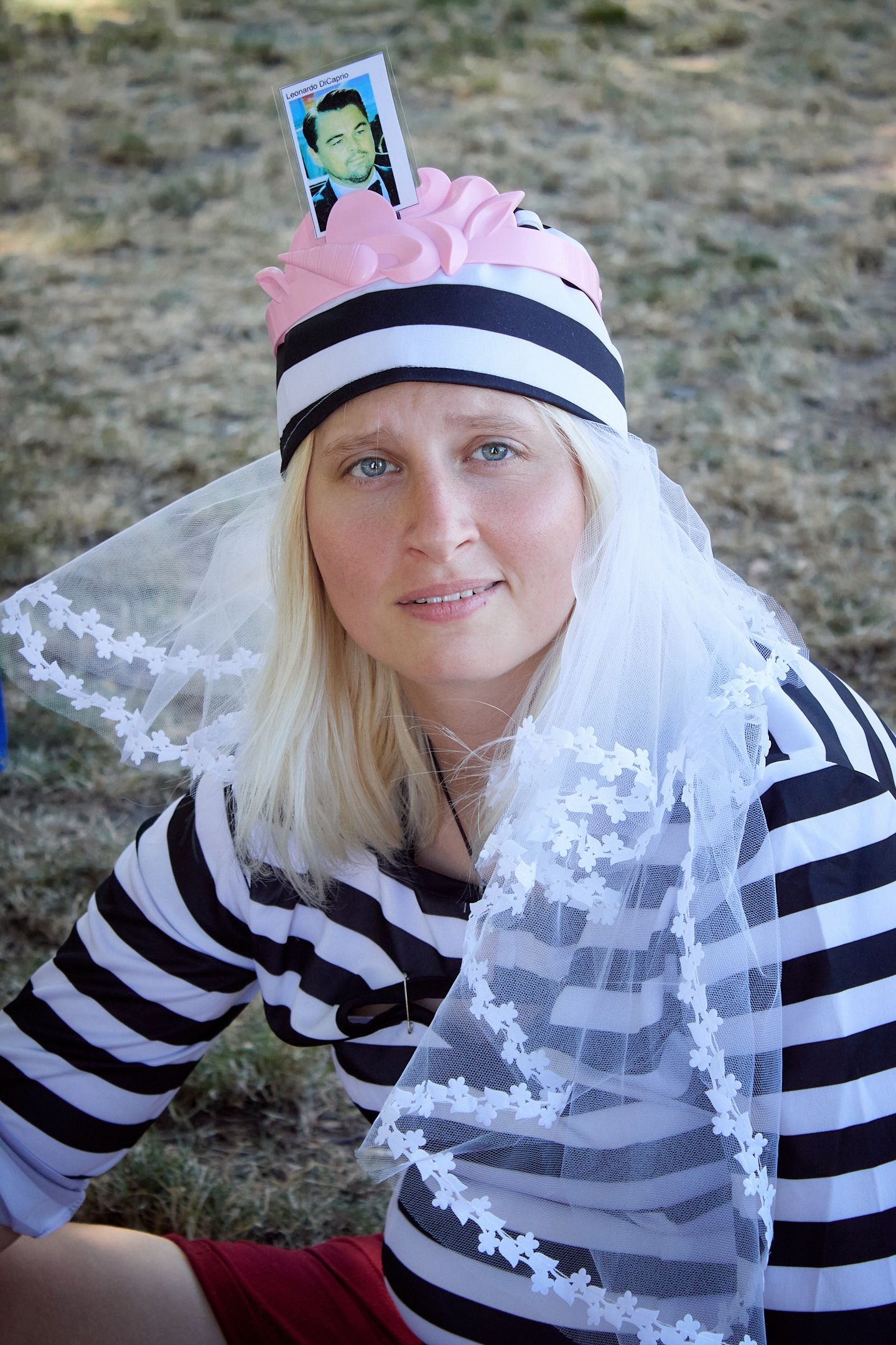 Blonde woman in striped outfit wearing decorative pink hat with photo and white lace veil outdoors.