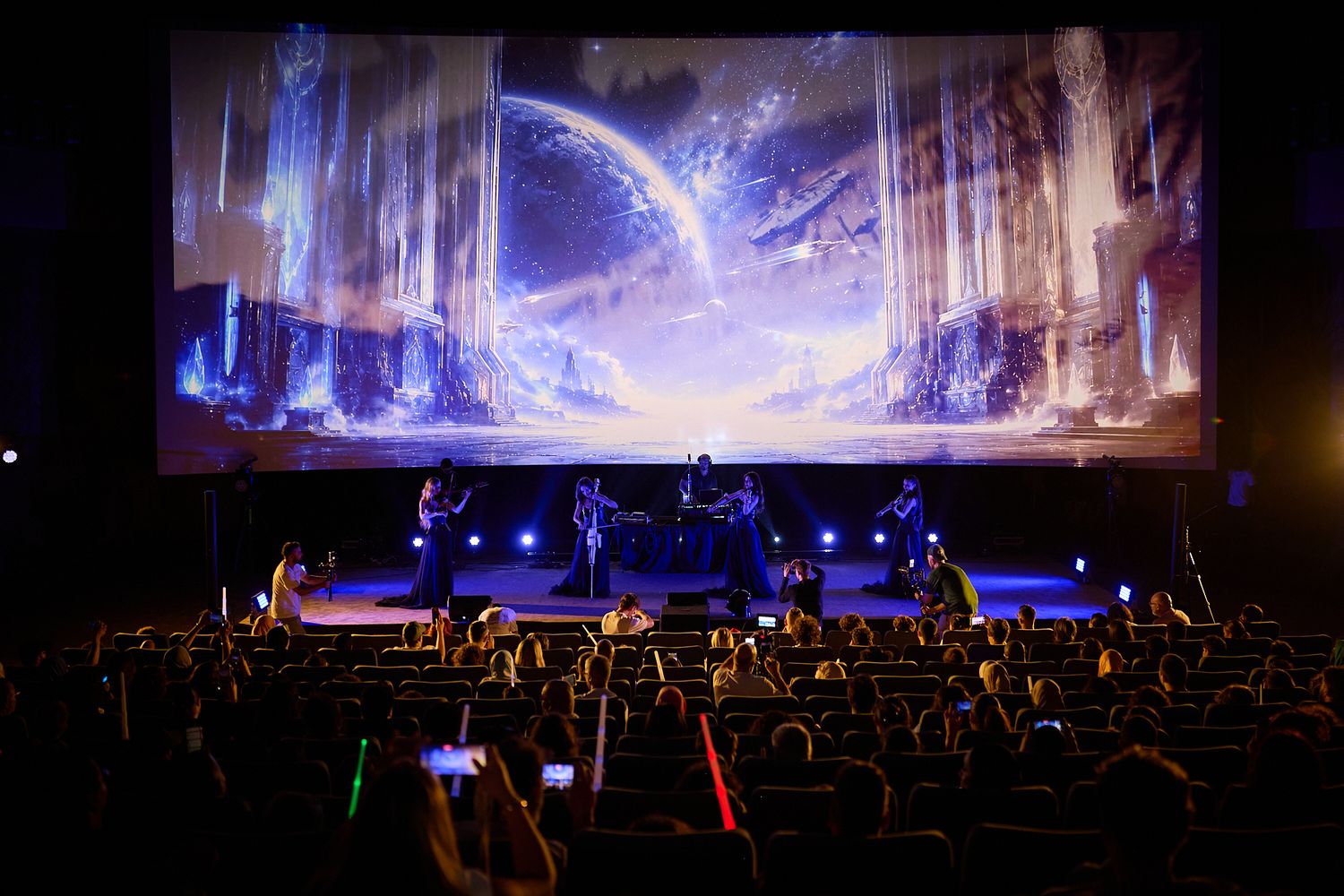 Live concert with band performing on stage beneath a stunning fantasy-themed projection screen in a dark theater.