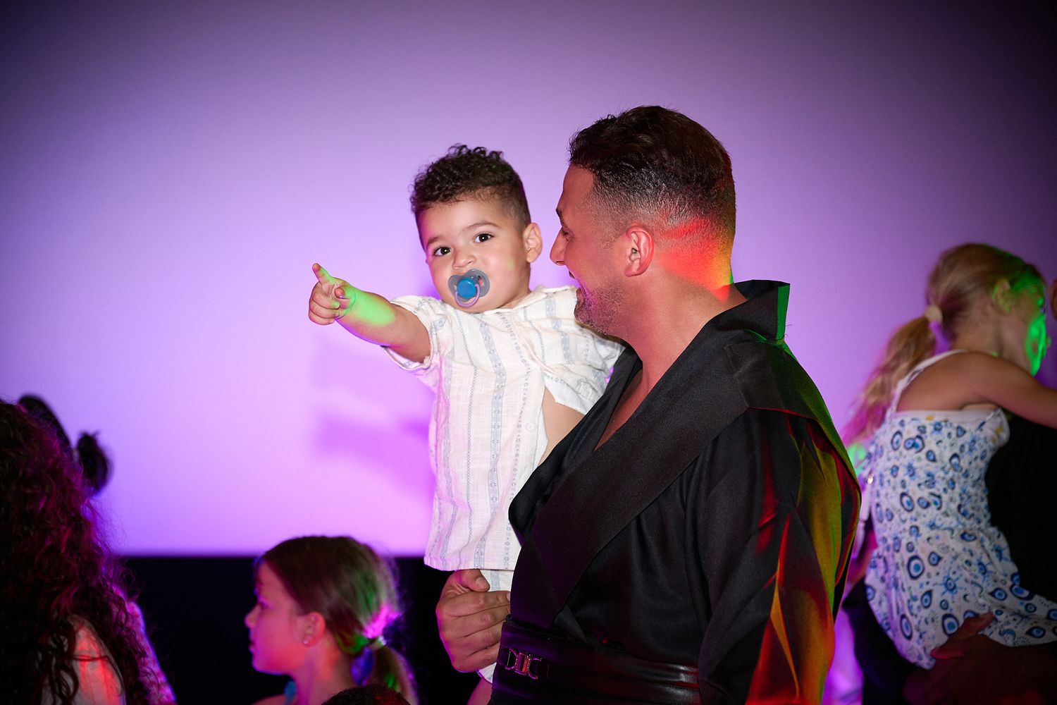 A man holds a baby with a blue pacifier at a colorful party, as the child points toward something nearby.