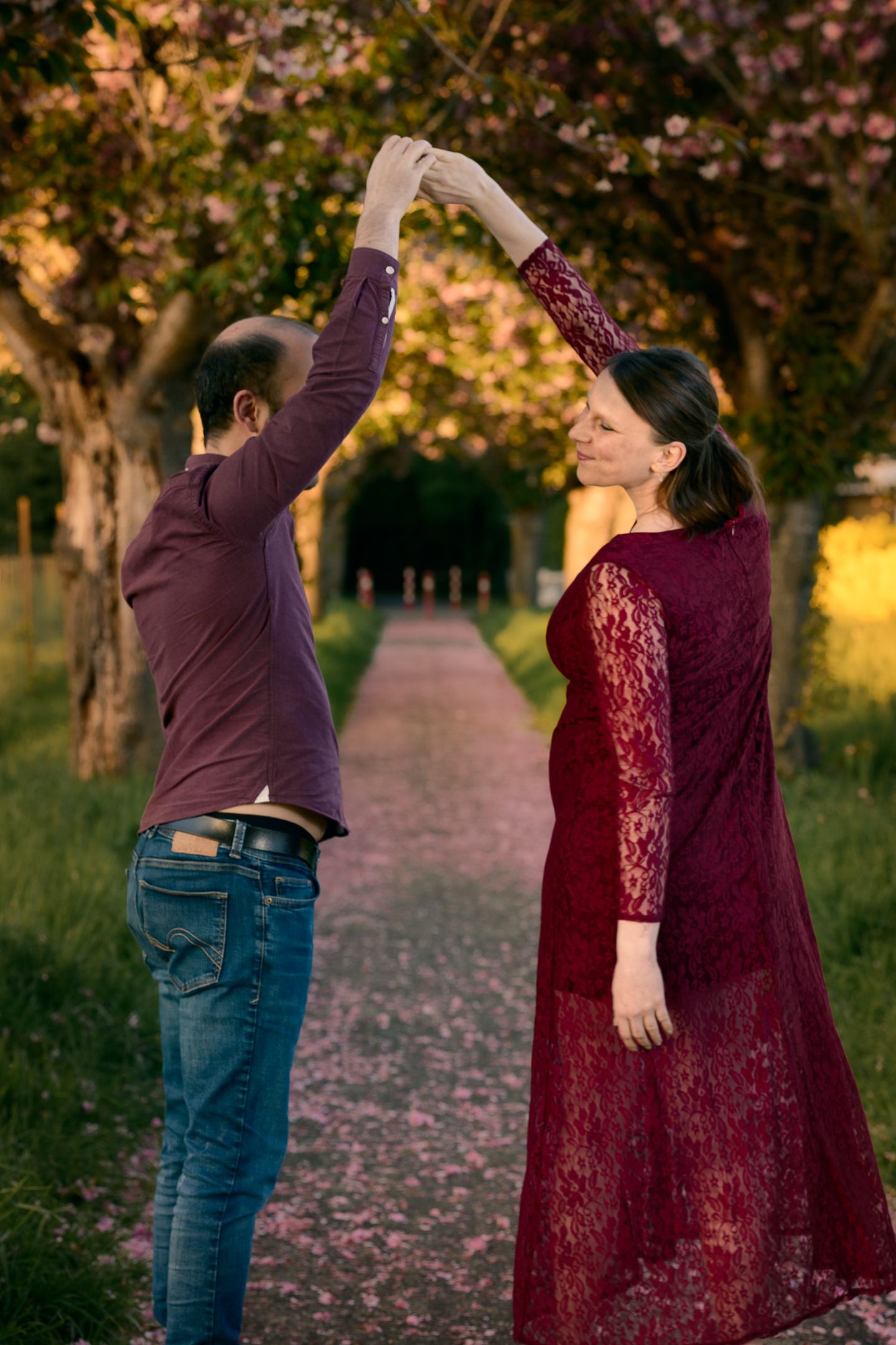 Couple dancing outdoors among blooming trees at sunset, woman in burgundy lace dress being twirled by man in jeans.