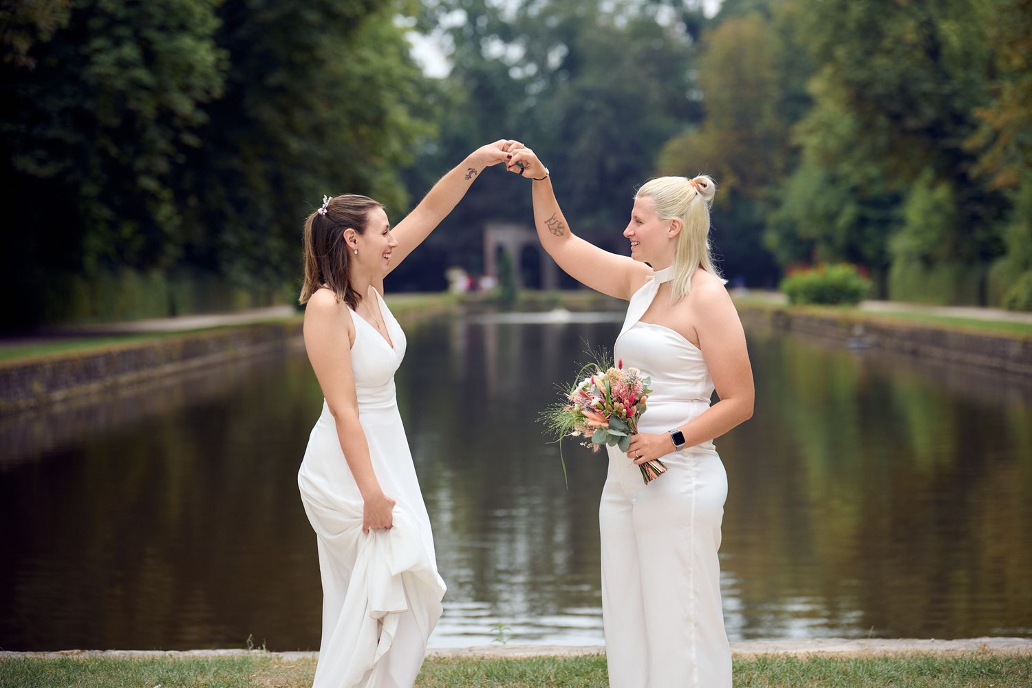 Two brides in white dresses dancing by a scenic canal in a park on their wedding day.