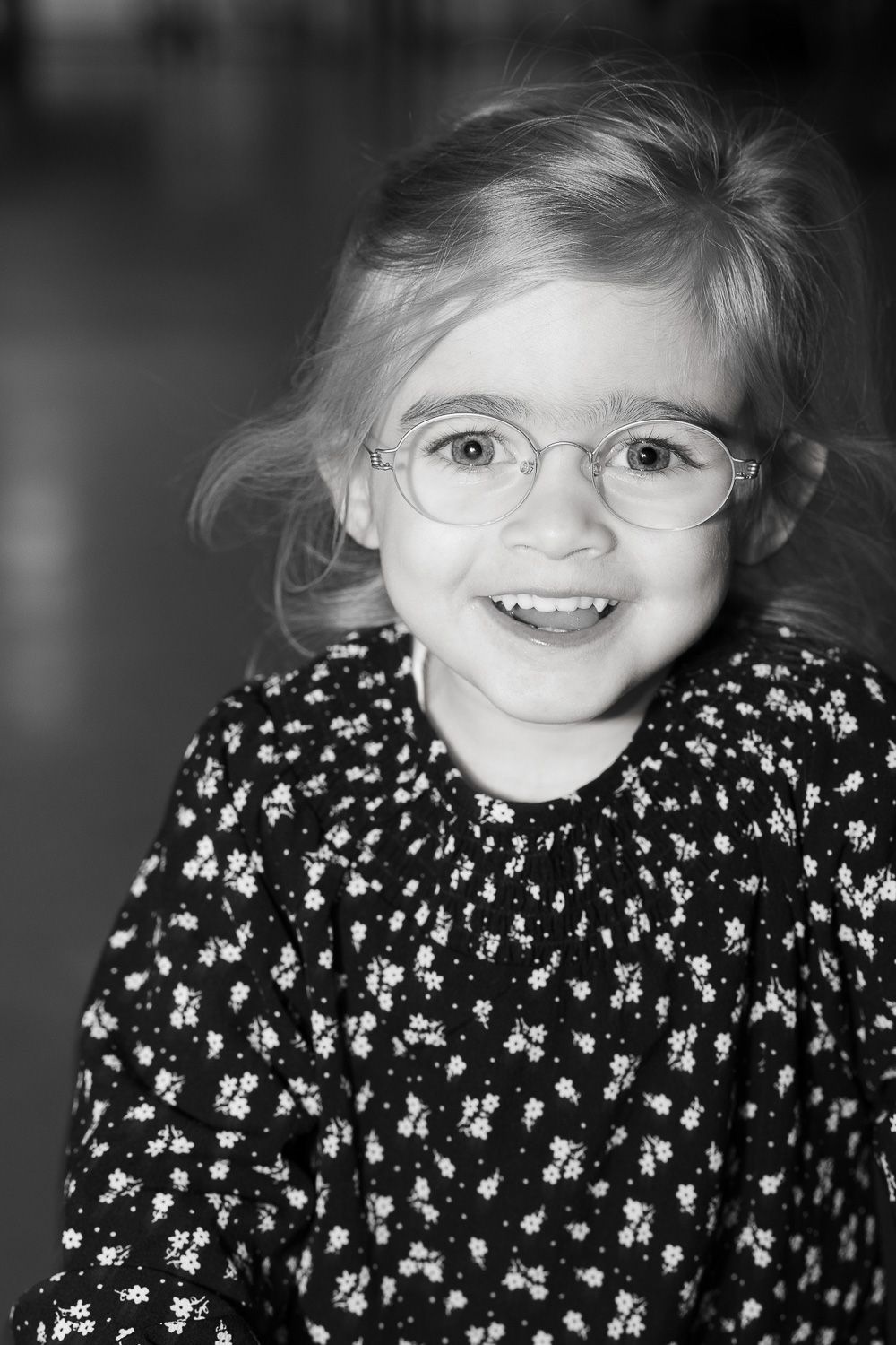 Black and white portrait of a smiling young blonde girl wearing round glasses and a floral dress.