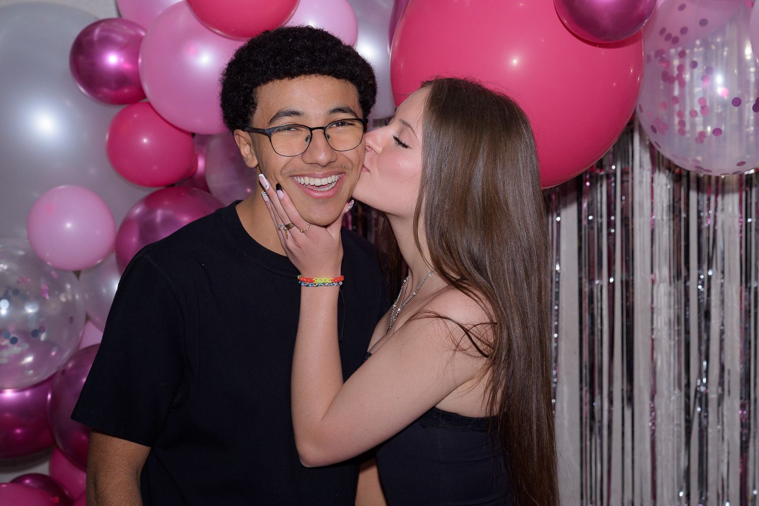 A young couple poses in front of pink and white balloon backdrop, girl kissing boy on cheek at party.