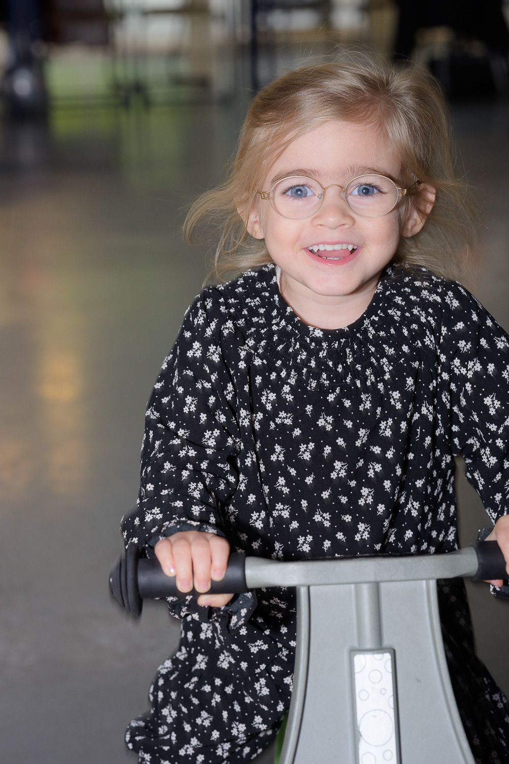 Adorable toddler girl with glasses smiling while riding a balance bike indoors.