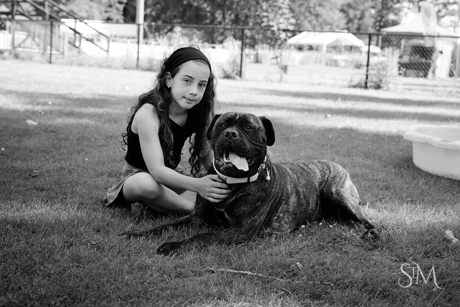 Black and white photo of a young girl sitting on grass with a large Mastiff dog in a park setting.