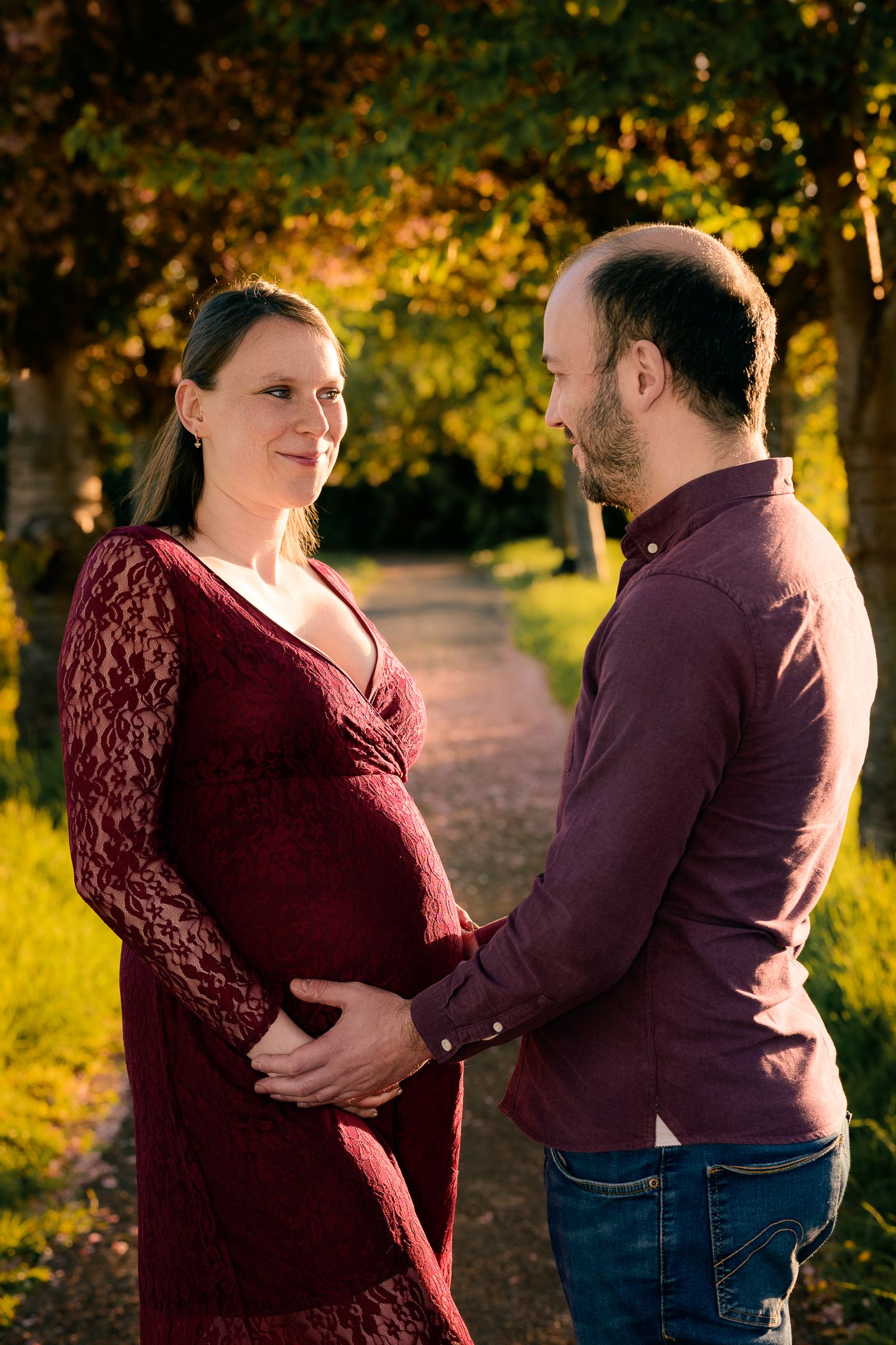 Pregnant woman in red lace dress with partner cradling baby bump outdoors during golden hour autumn maternity photoshoot.