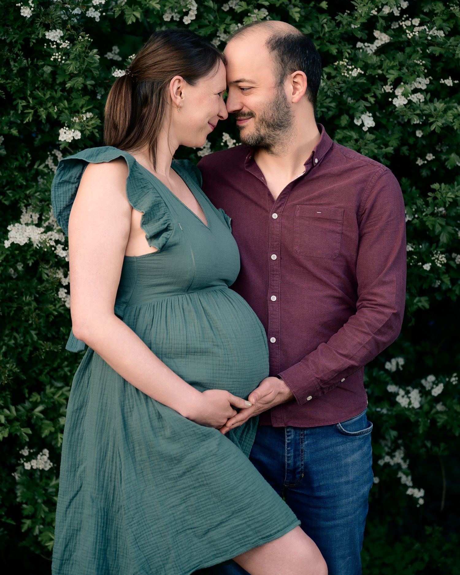 Pregnant couple embracing outdoors near white flowering bushes, woman in green dress, man in burgundy shirt and jeans.