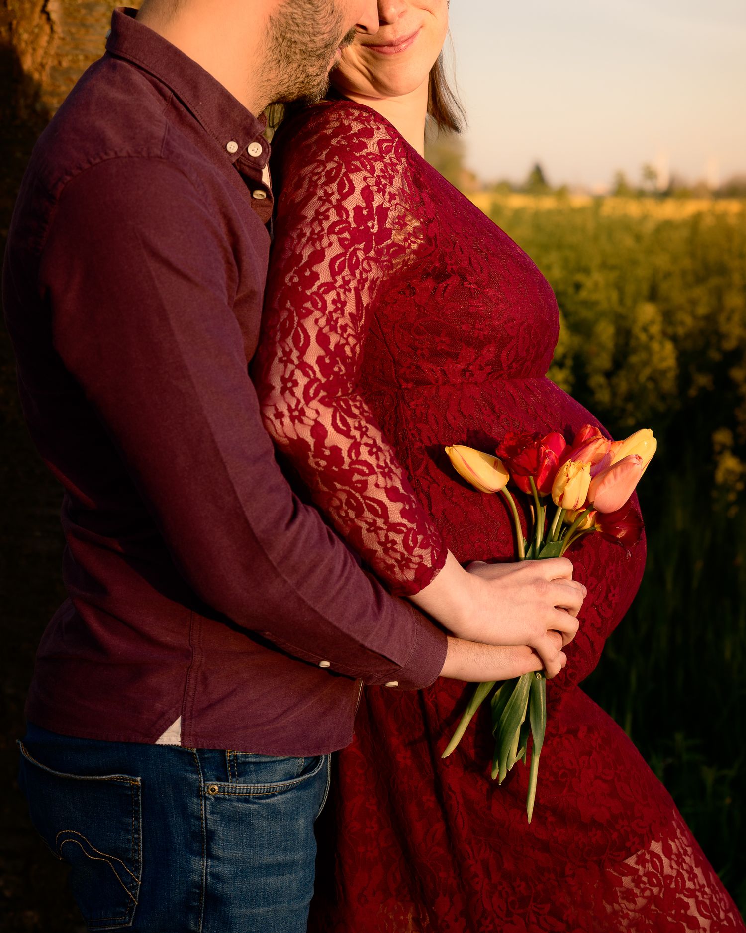 Couple embracing outdoors, woman in red lace dress holding colorful tulips, golden hour light.