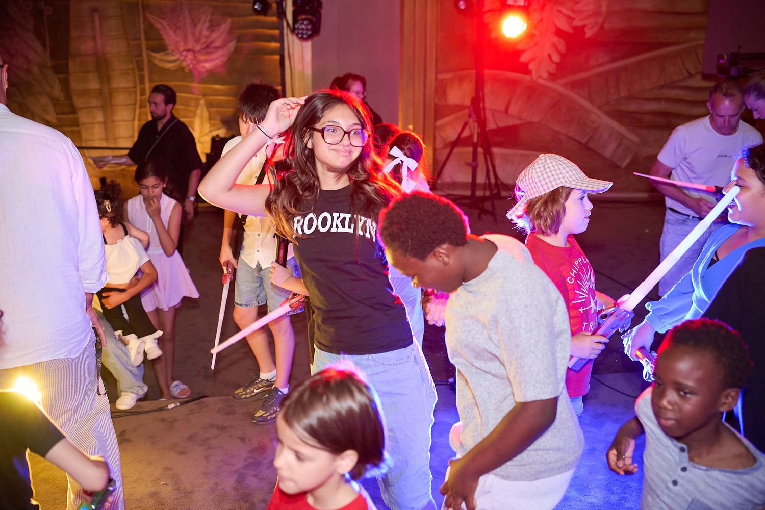 Children dancing at a lively indoor party with colorful lights, one girl wearing a Brooklyn t-shirt.
