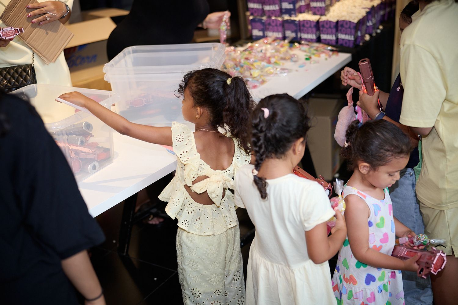 Young girls in white dresses reaching into a clear bin at a colorful candy or craft table at a party.