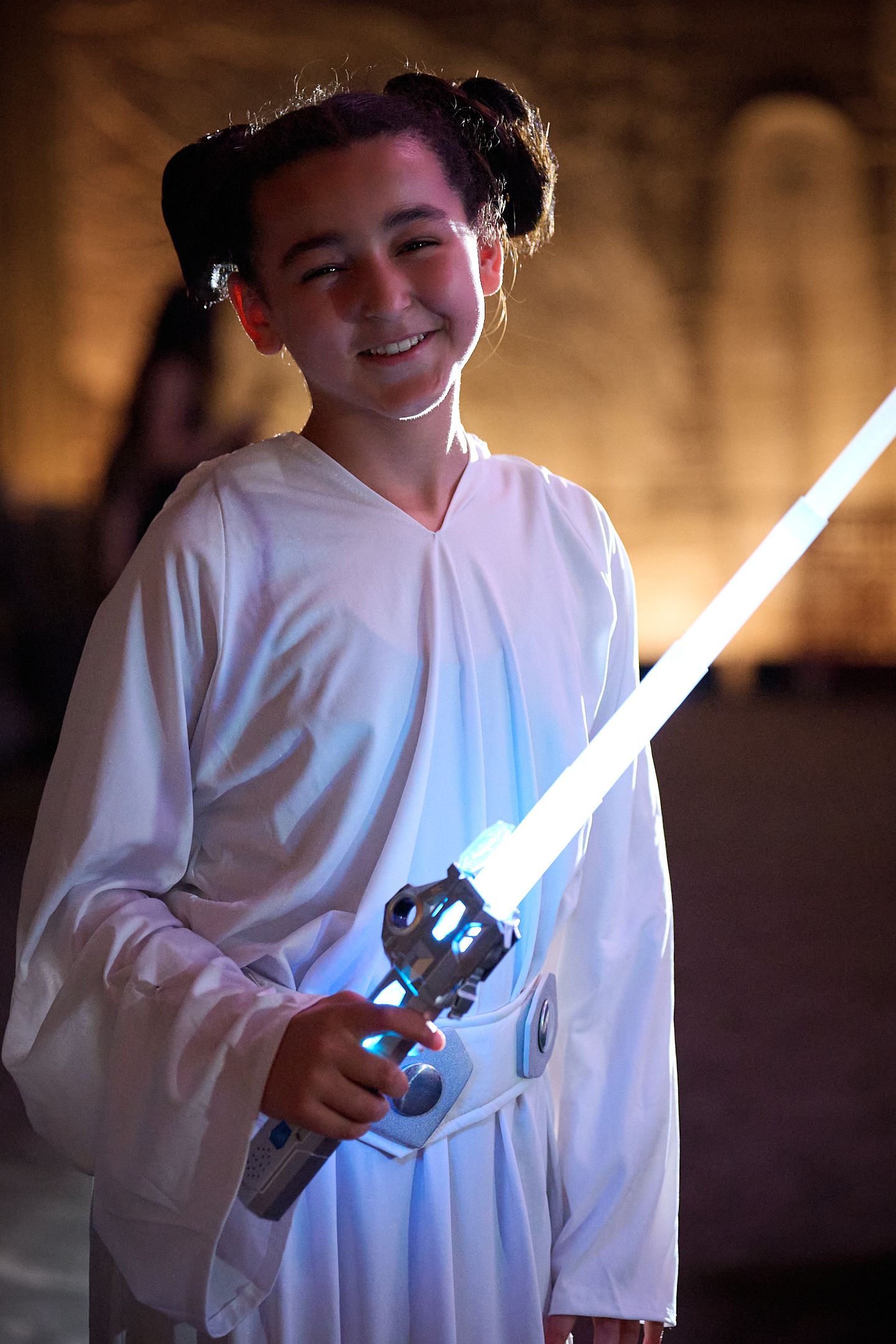Child dressed as Princess Leia in white costume holding glowing lightsaber toy, smiling at camera.