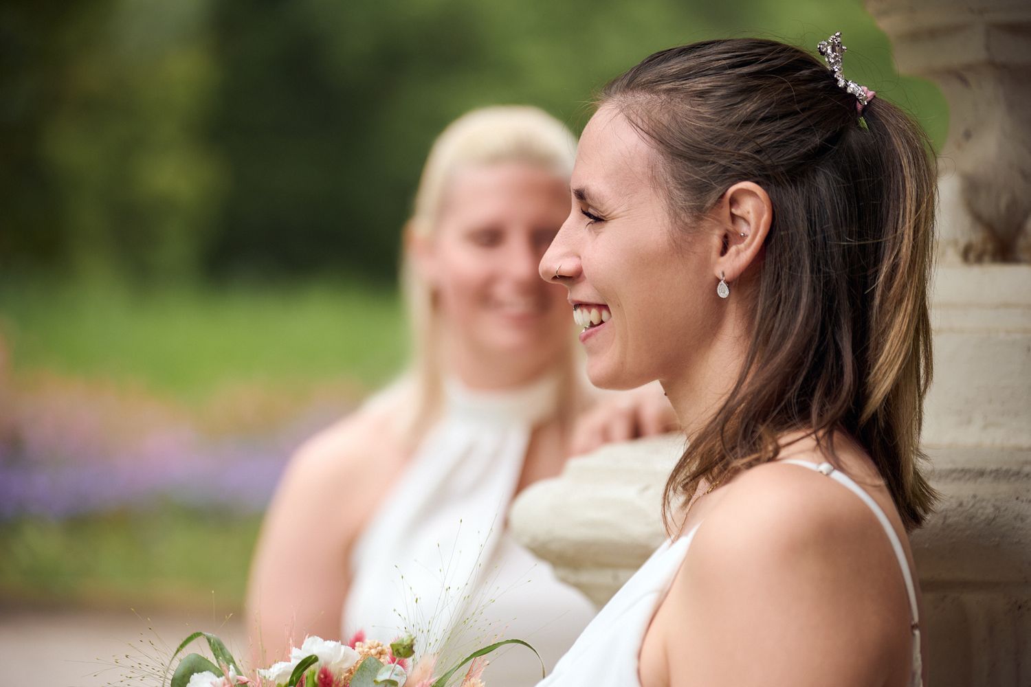 Two brides in white dresses laugh together outdoors, one holding a bouquet, with a stone fountain behind them.