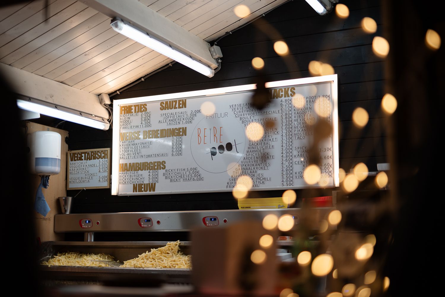 Cozy food stall menu board illuminated by warm string lights and fluorescent lighting in a rustic wooden kiosk.
