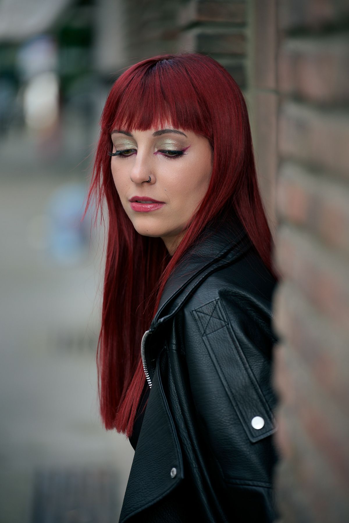A woman with vibrant red hair and bangs wearing a black leather jacket, posing against a brick wall outdoors.