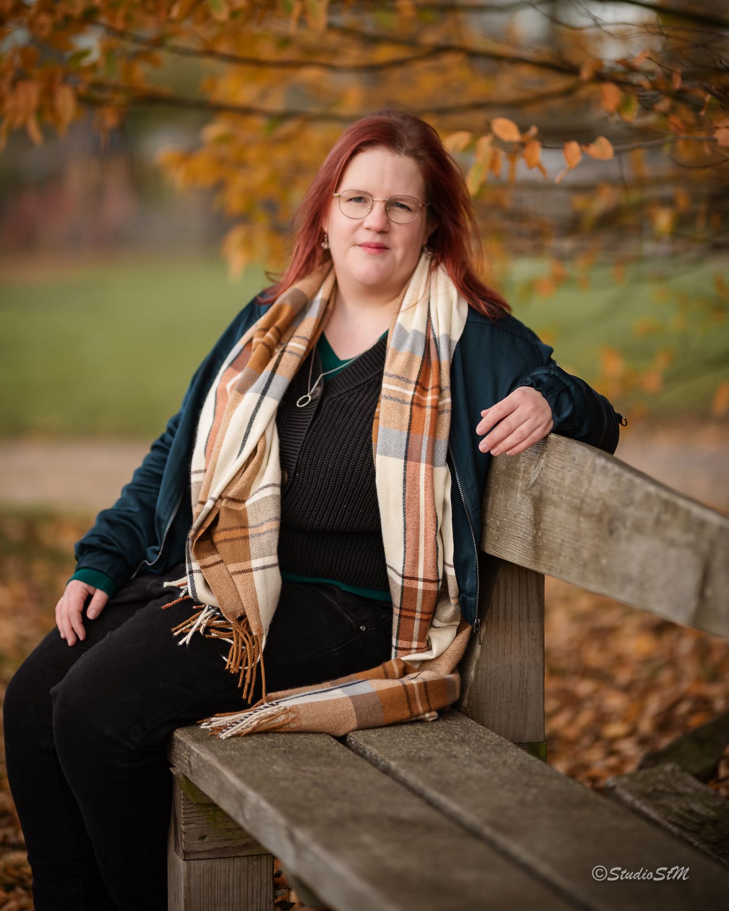 Red-haired woman in teal jacket and plaid scarf sitting on wooden bench under autumn foliage.