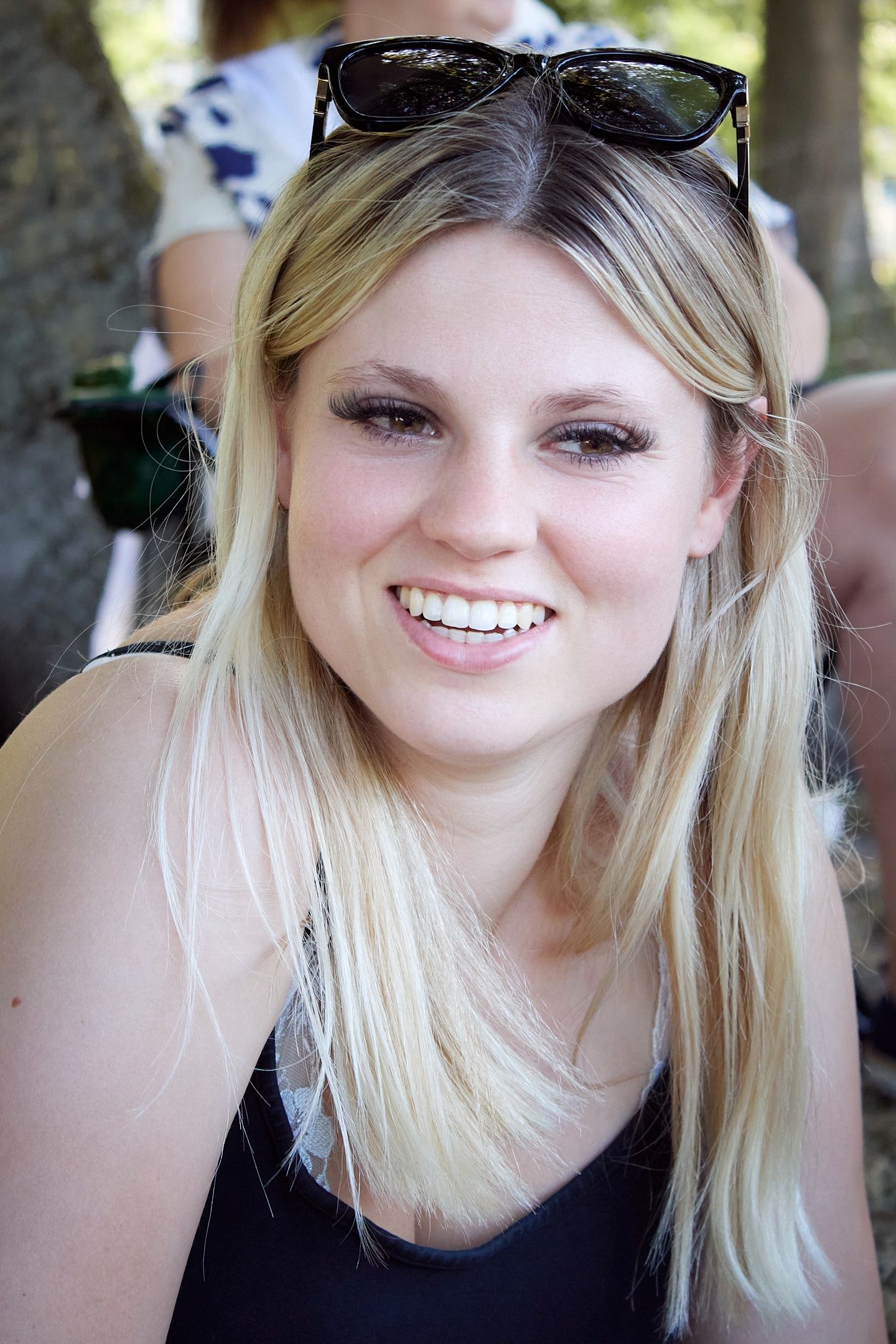 Smiling blonde woman with sunglasses on her head wearing a black top outdoors.
