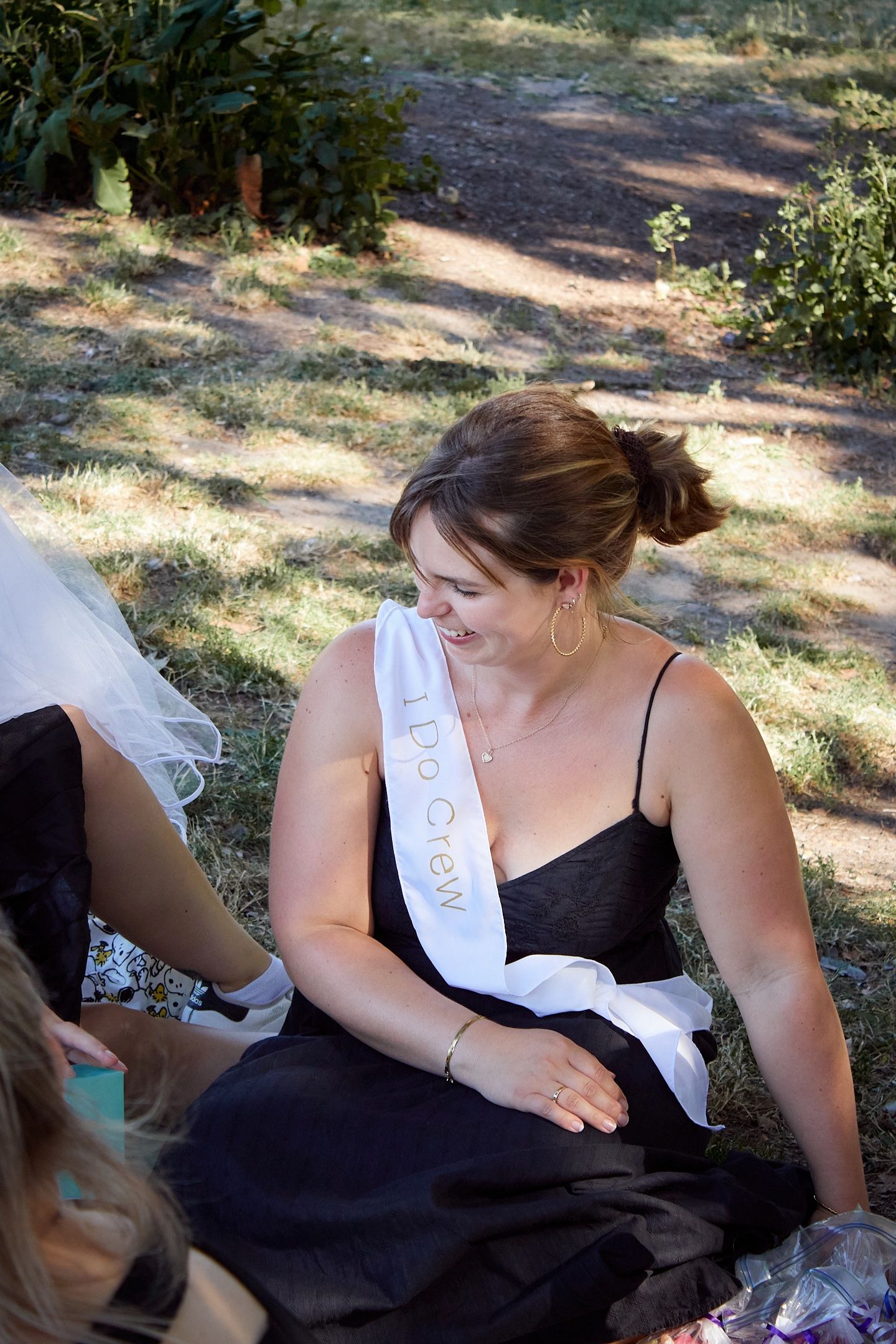 Woman in black dress wearing a white 'I Do Crew' sash sits outdoors laughing at a bachelorette party.