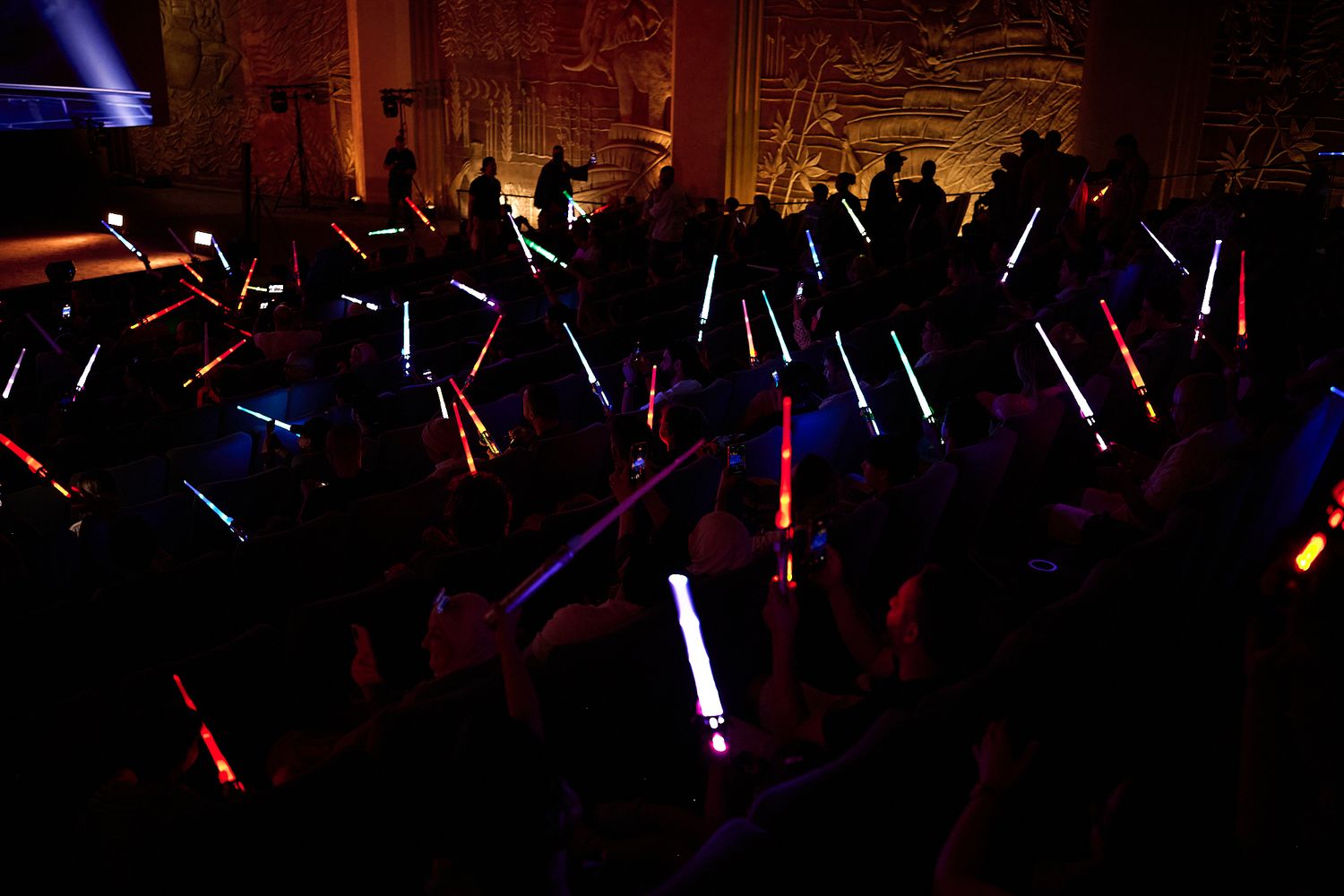 Crowd holding colorful glowing lightsabers in a dark venue during a Star Wars themed event.