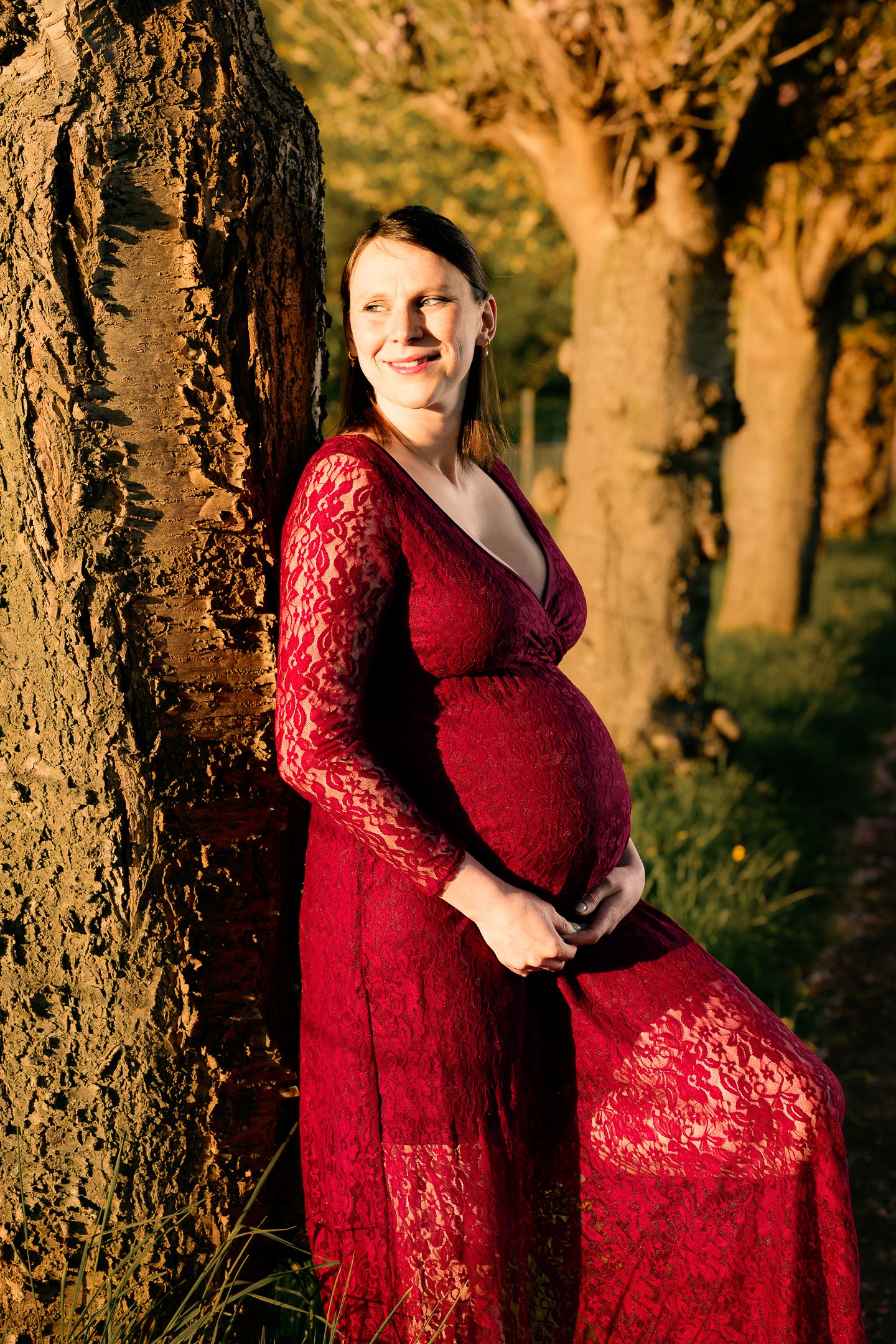 Smiling pregnant woman in red lace dress leaning against tree in golden sunlight park setting.