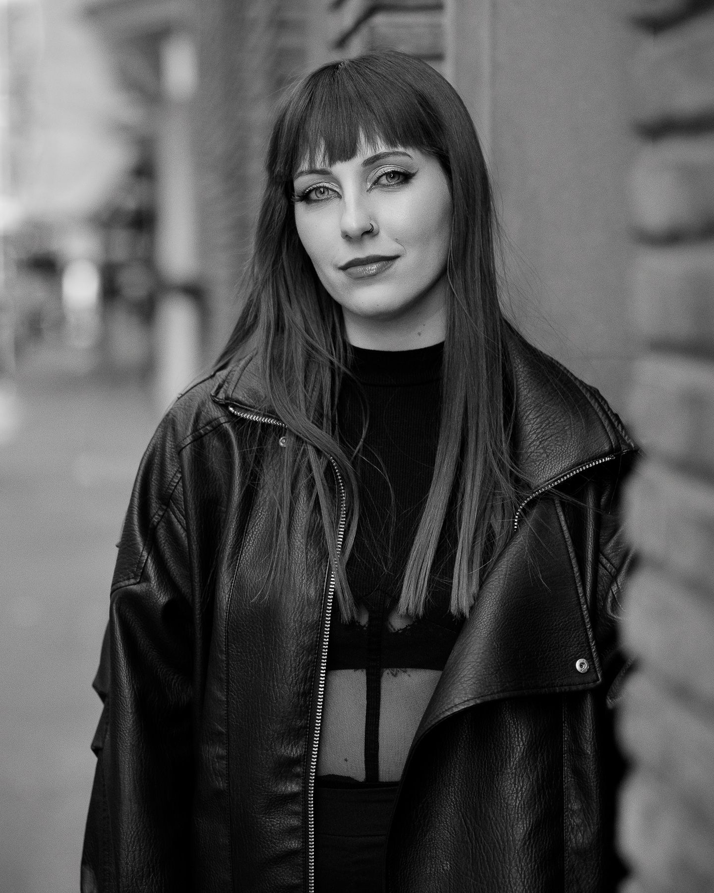 Black and white portrait of a young woman with bangs wearing an oversized leather jacket on a city street.