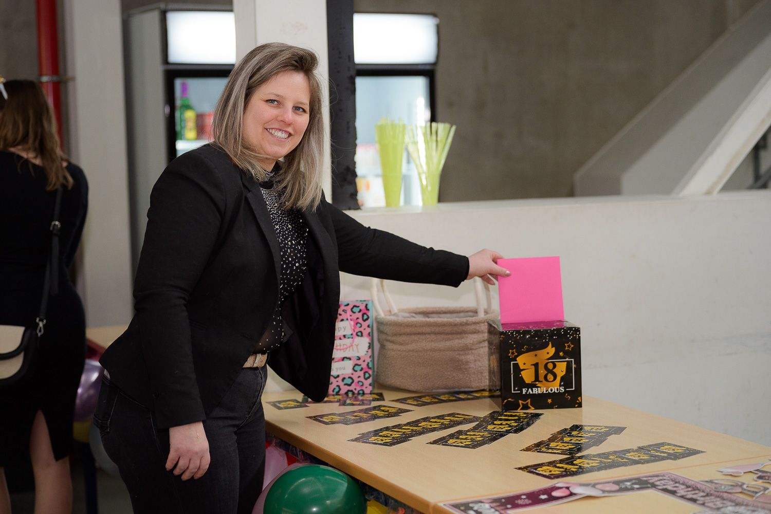 Smiling woman in black blazer placing a pink card into a decorated gift box at a celebration table.