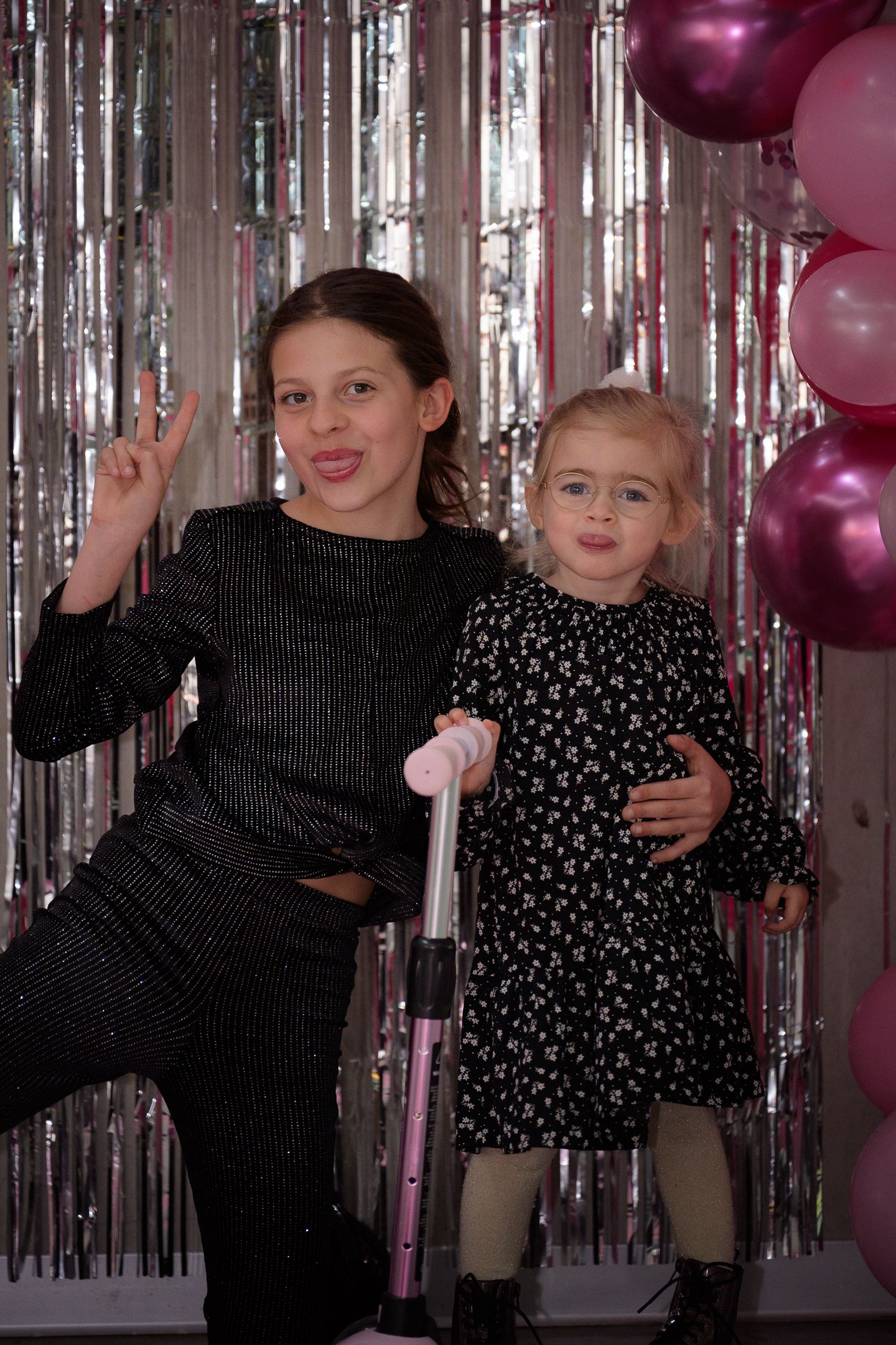 Two girls posing in front of silver tinsel curtain with pink balloons at a birthday party.
