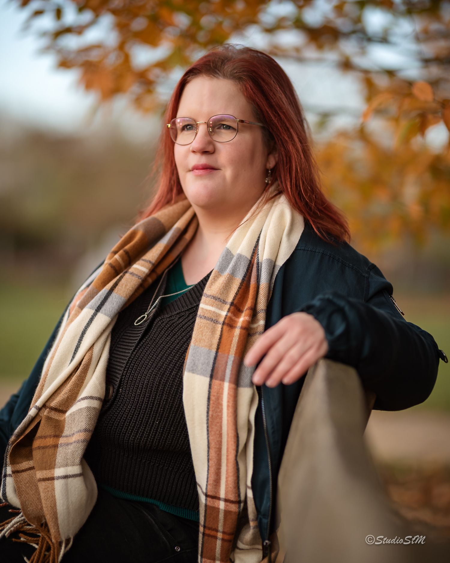 Red-haired woman in teal jacket and plaid scarf posing under golden autumn leaves in park.