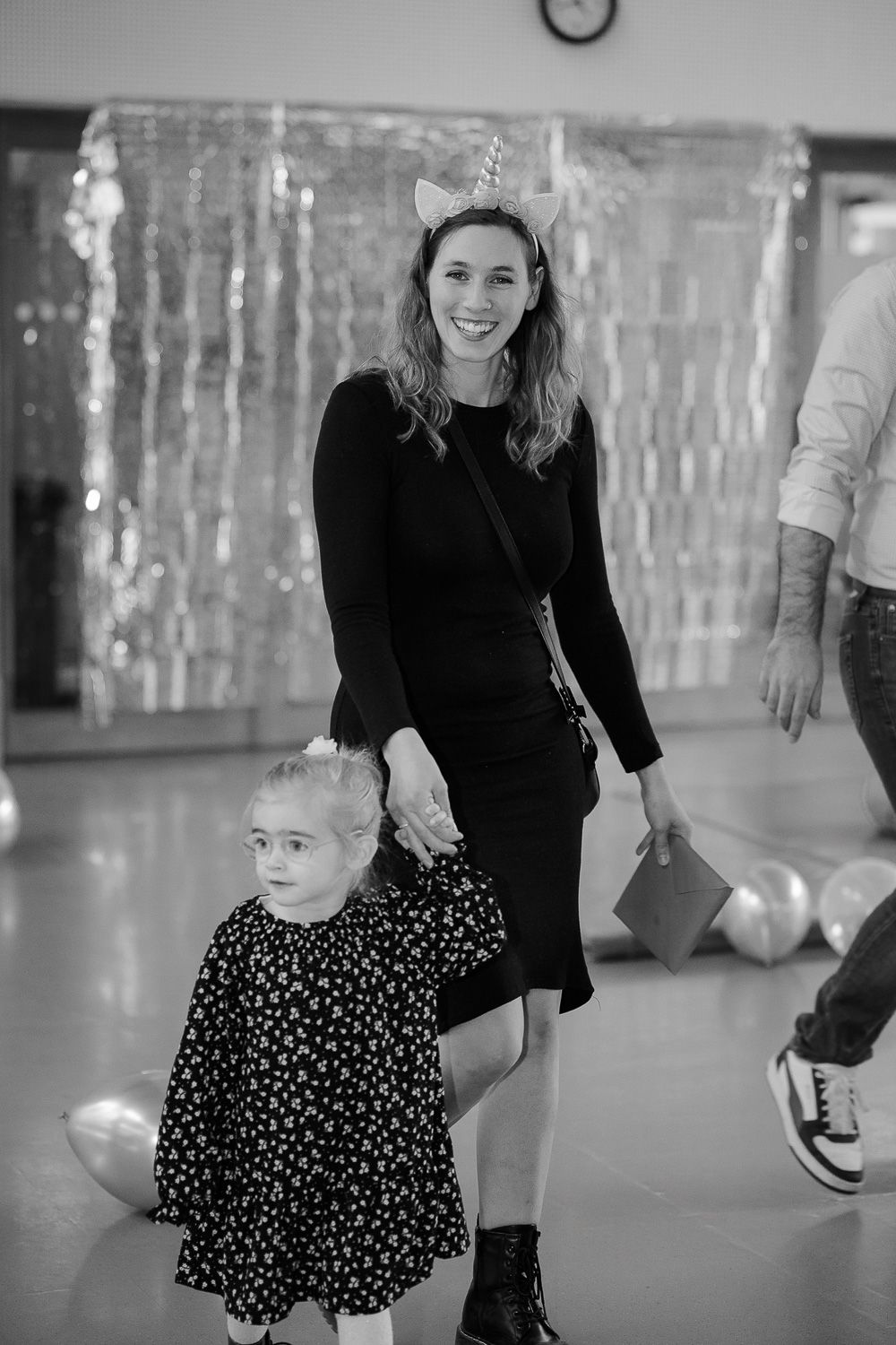 Woman in black dress with unicorn headband holds young girl's hand at a festive party with balloons.