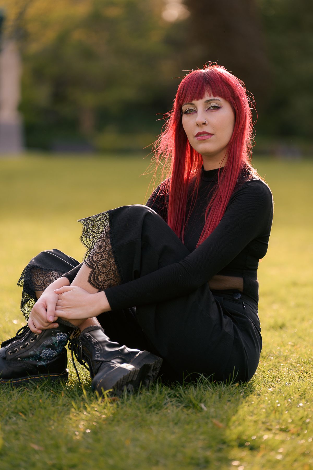 Young woman with red hair in all-black outfit sitting on green grass in a park, wearing black boots.