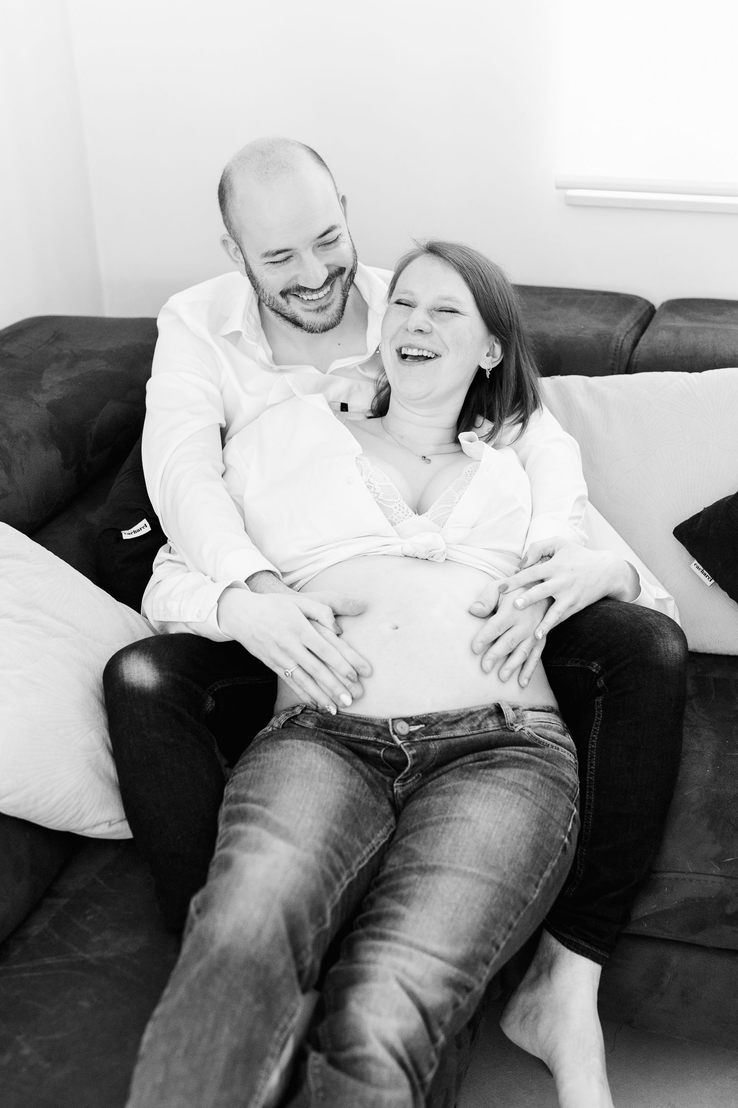 Black and white photo of a happy couple on a sofa, man embracing pregnant woman's belly, both laughing joyfully.