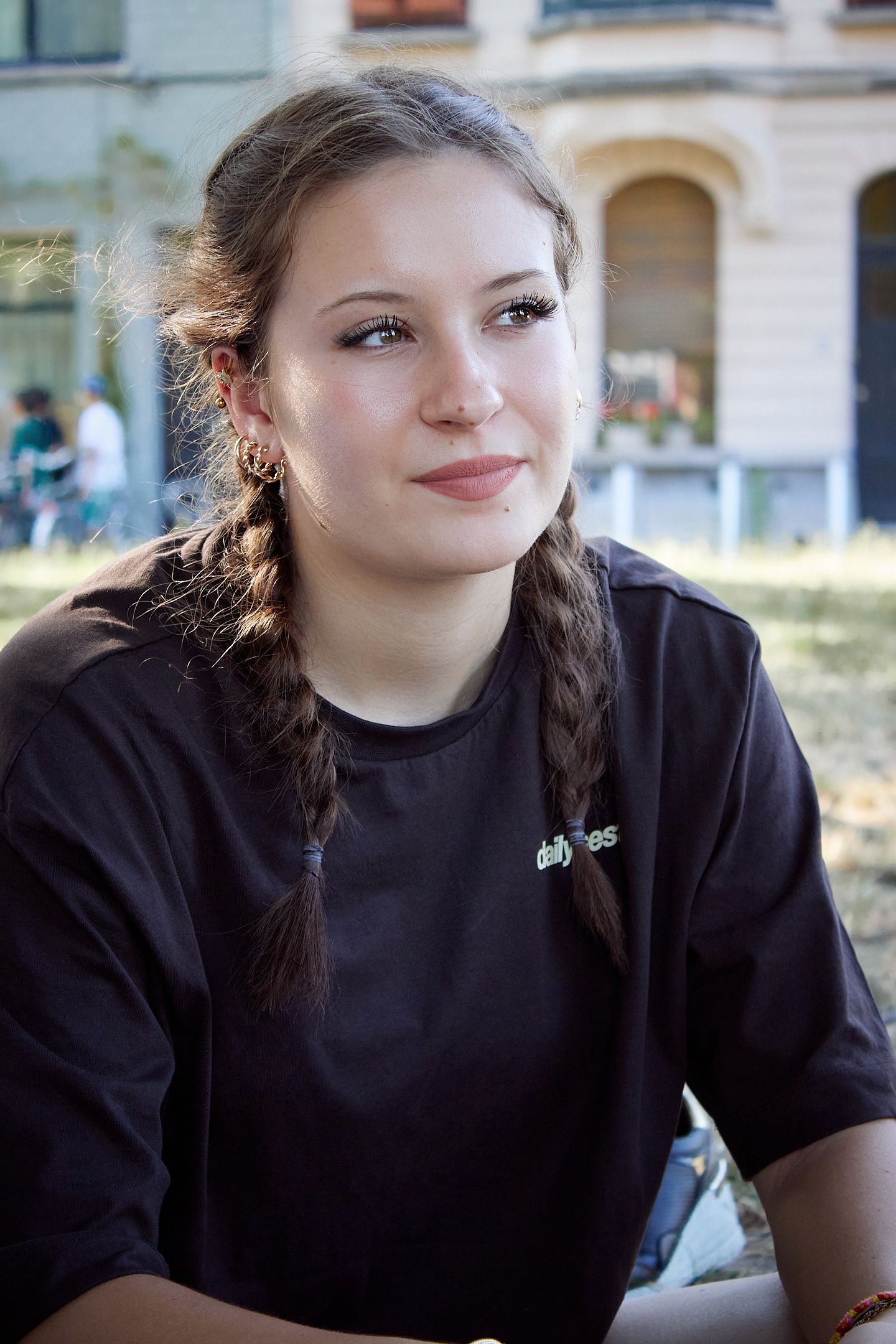 Young woman with braided pigtails wearing a black t-shirt, smiling outdoors with a building in the background.